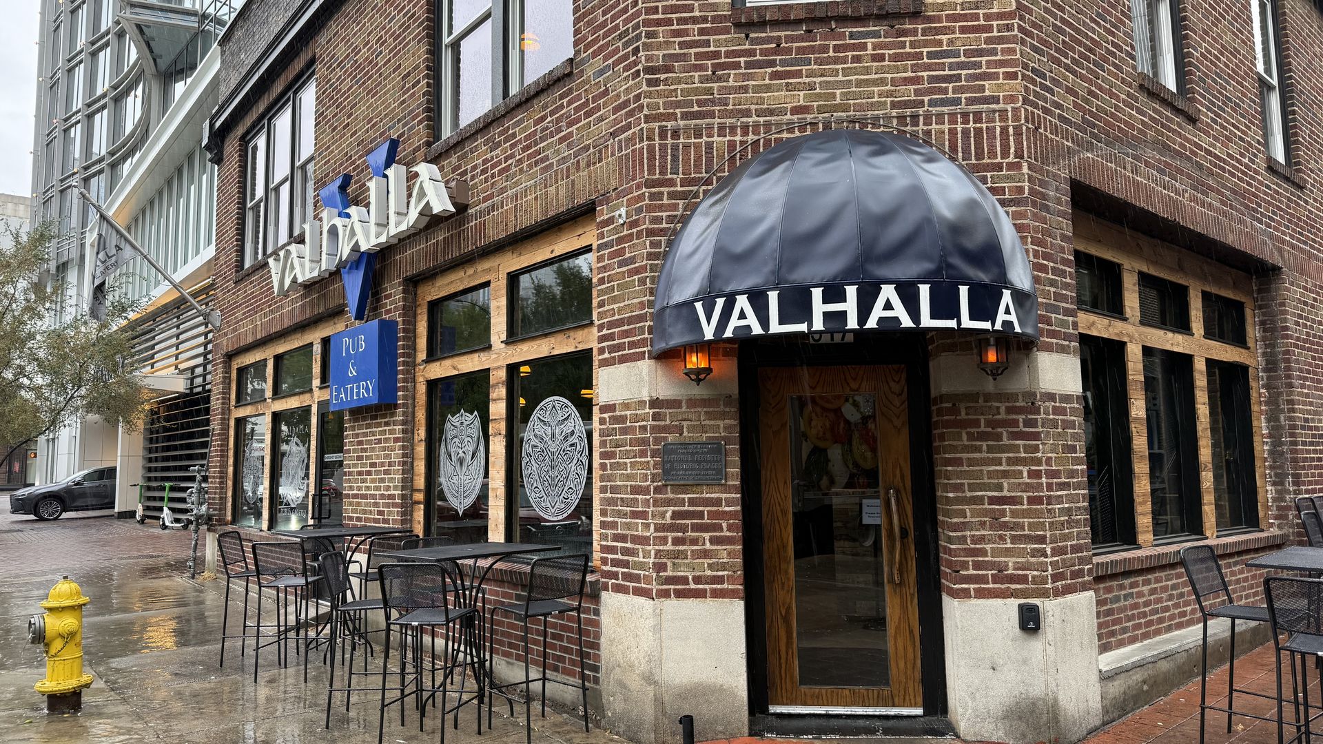 Corner view of Valhalla pub and eatery in a brick building with black metal tables and chairs outside on a wet sidewalk under a gray sky.