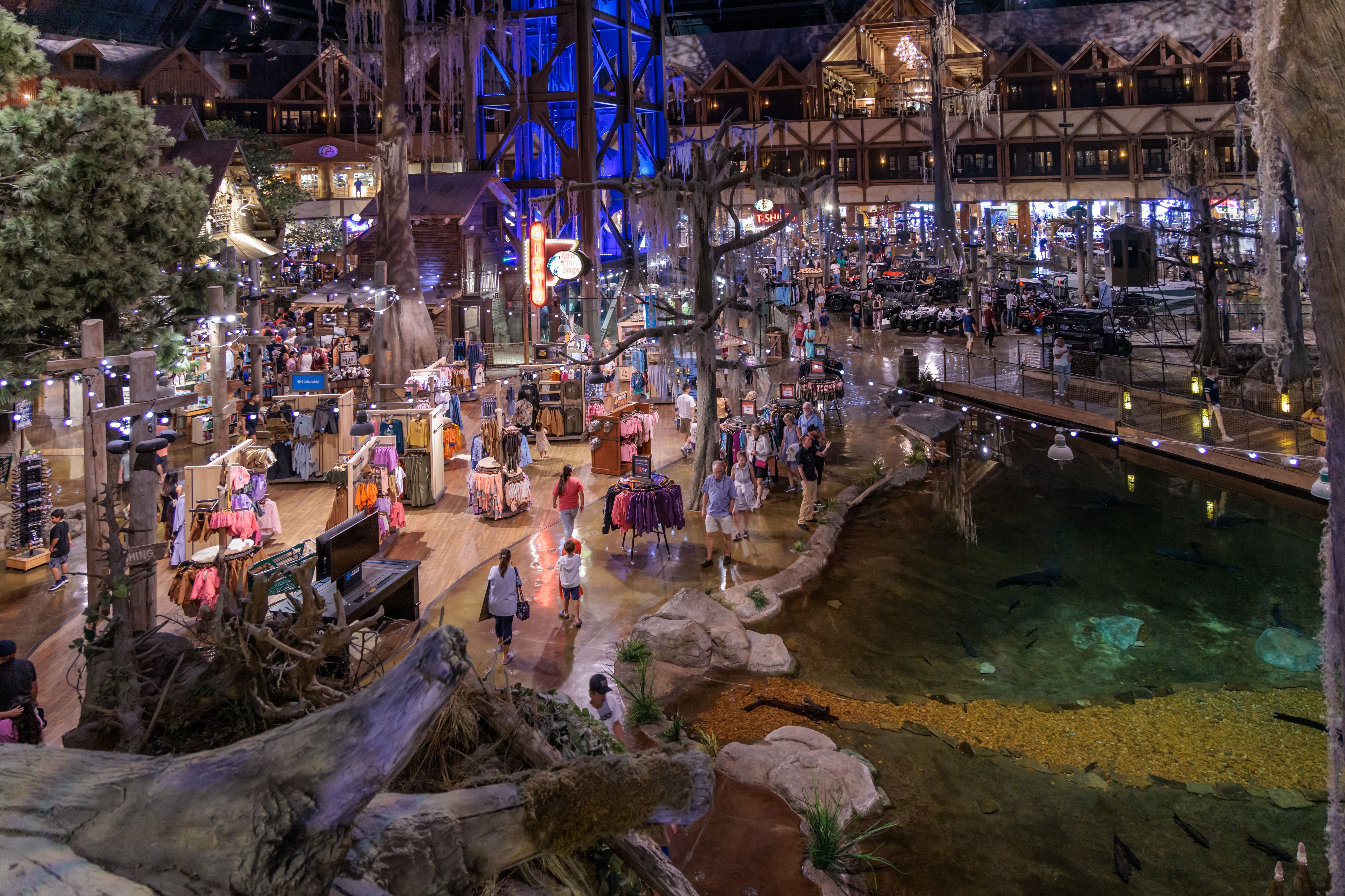 Indoor market hall with wooden stalls, shoppers browsing clothes and gifts, string lights, bare trees, a blue-lit central structure, and a rocky water feature along the edge.