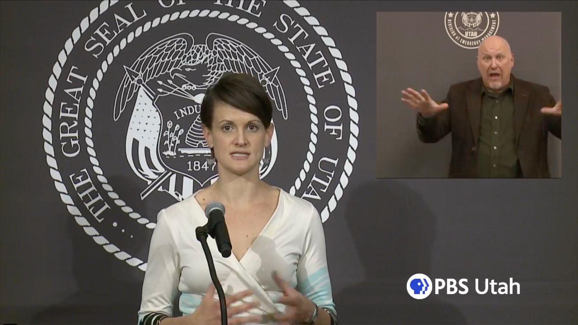 Dr Angela Dunn, state epidemiologist, speaks in front of a Utah state seal while an ASL interpreter translates in a chyron at  her right.
