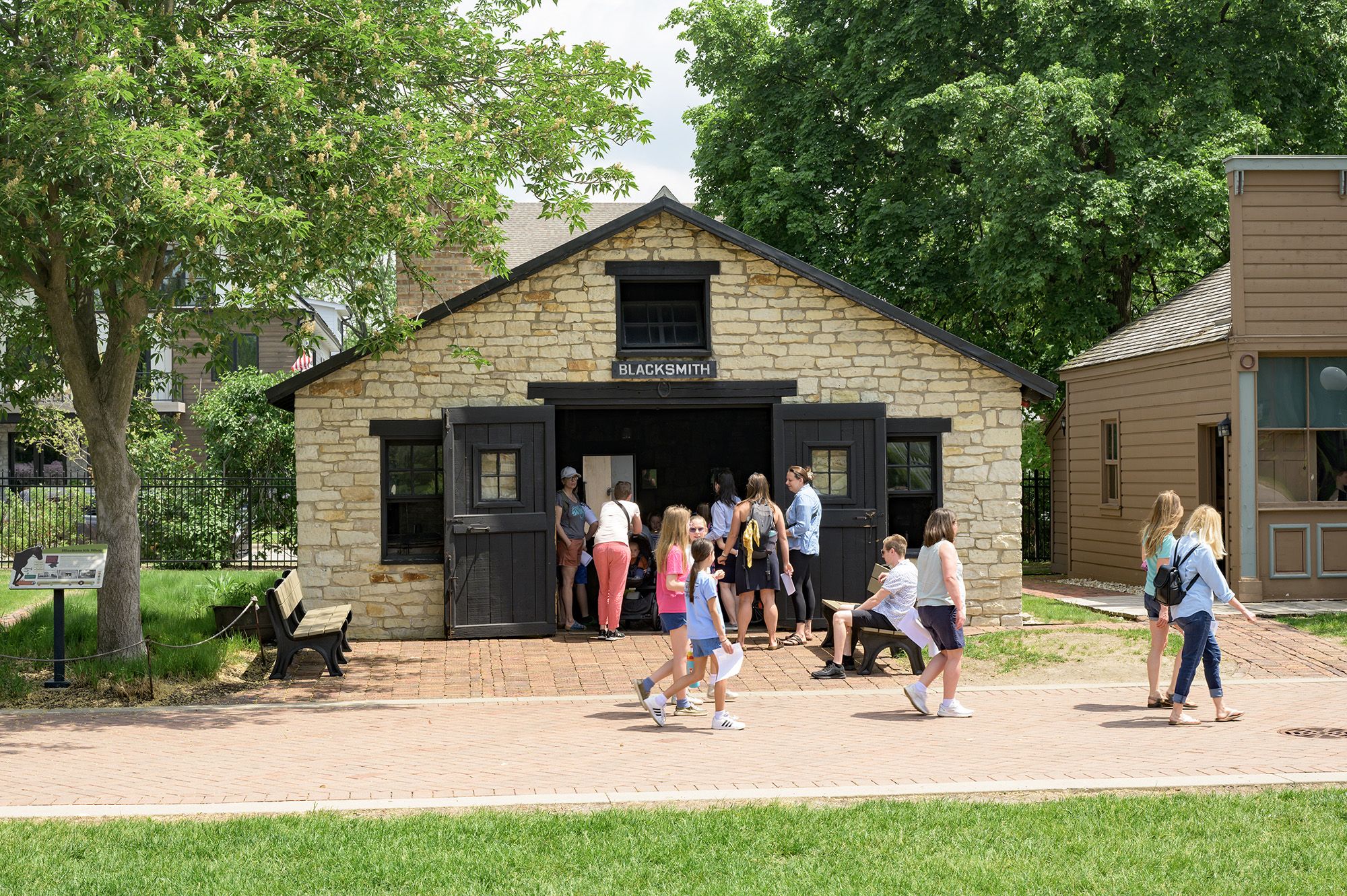 Stone building labeled BLACKSMITH with open black doors, people gathered around and walking nearby, trees and benches in a sunny outdoor museum or historical site.