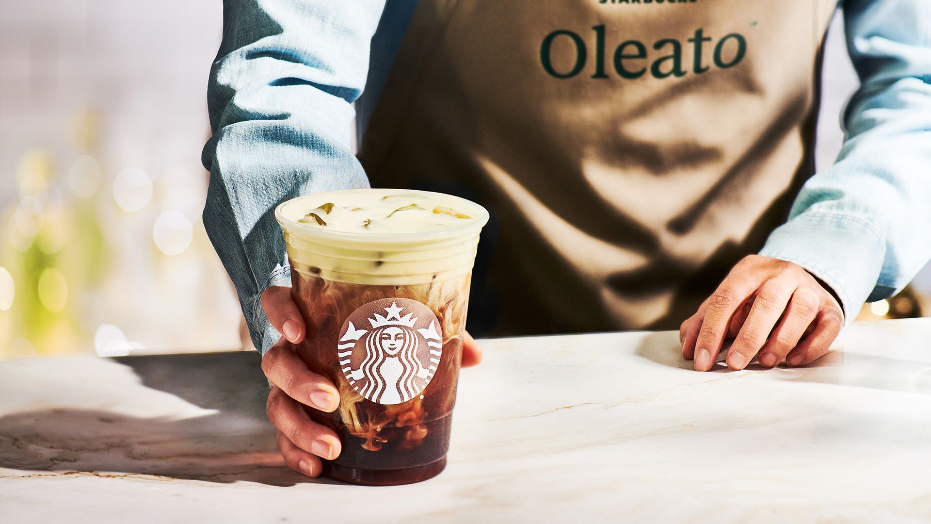 A person in an apron that says "Oleato" holding an iced coffee on a countertop.