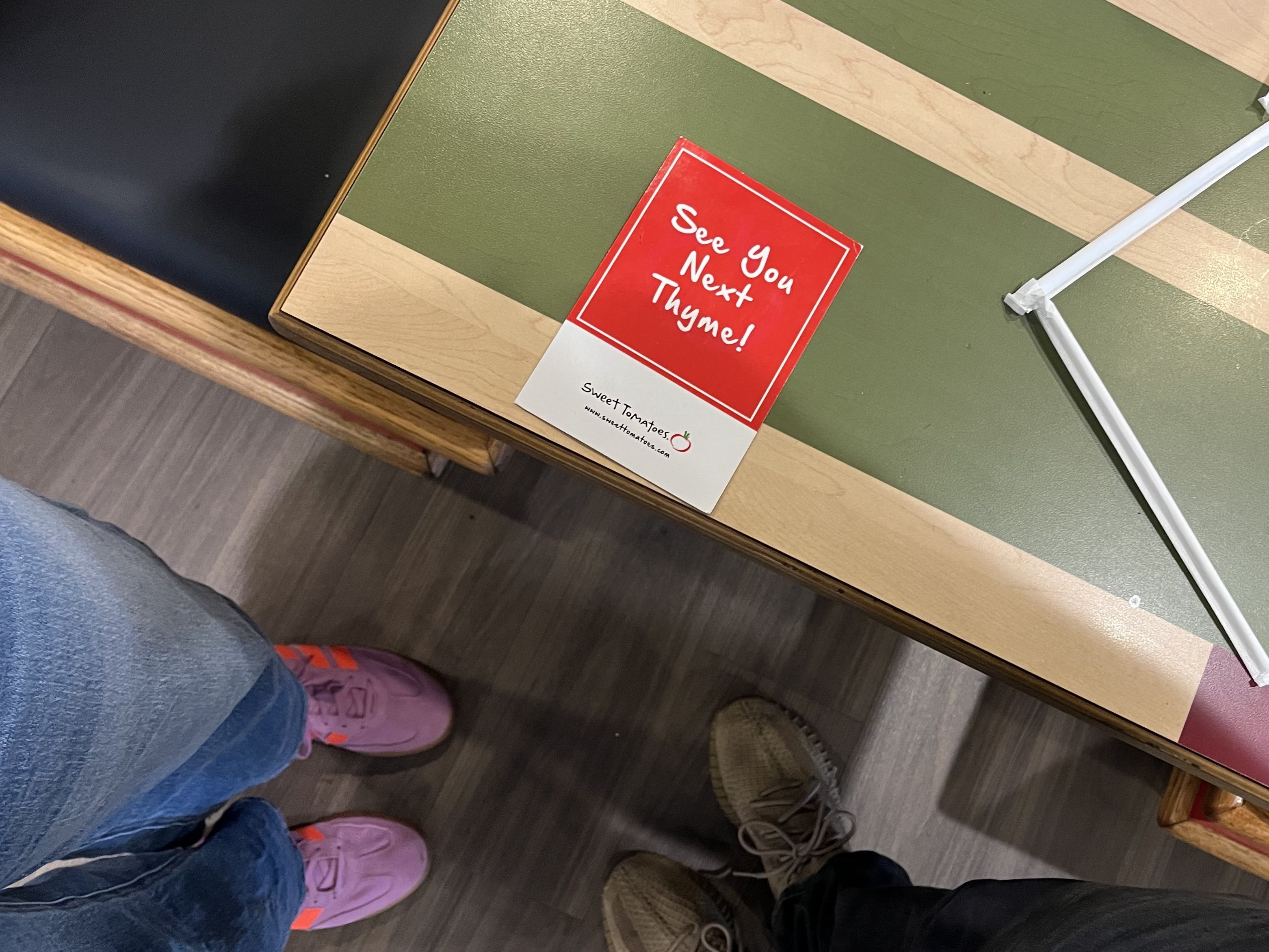 Red card on a green and beige striped table reads "See You Next Thyme!" in white script. A jeans-clad leg and pink shoe appear in the left foreground, with a white pole crossing the scene.