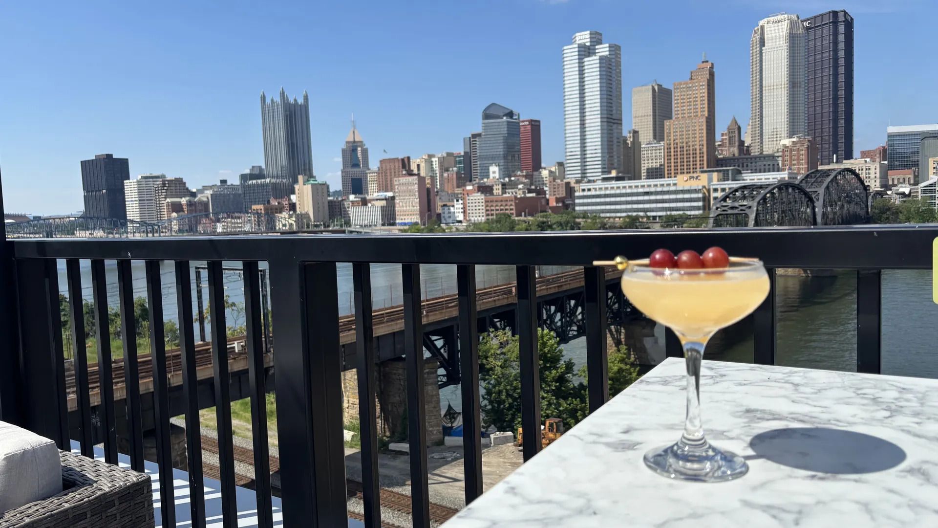 Clear blue sky over a city skyline across a wide river. A black railing and marble table frame a pale yellow cocktail with three red cherries on a skewer in the foreground.