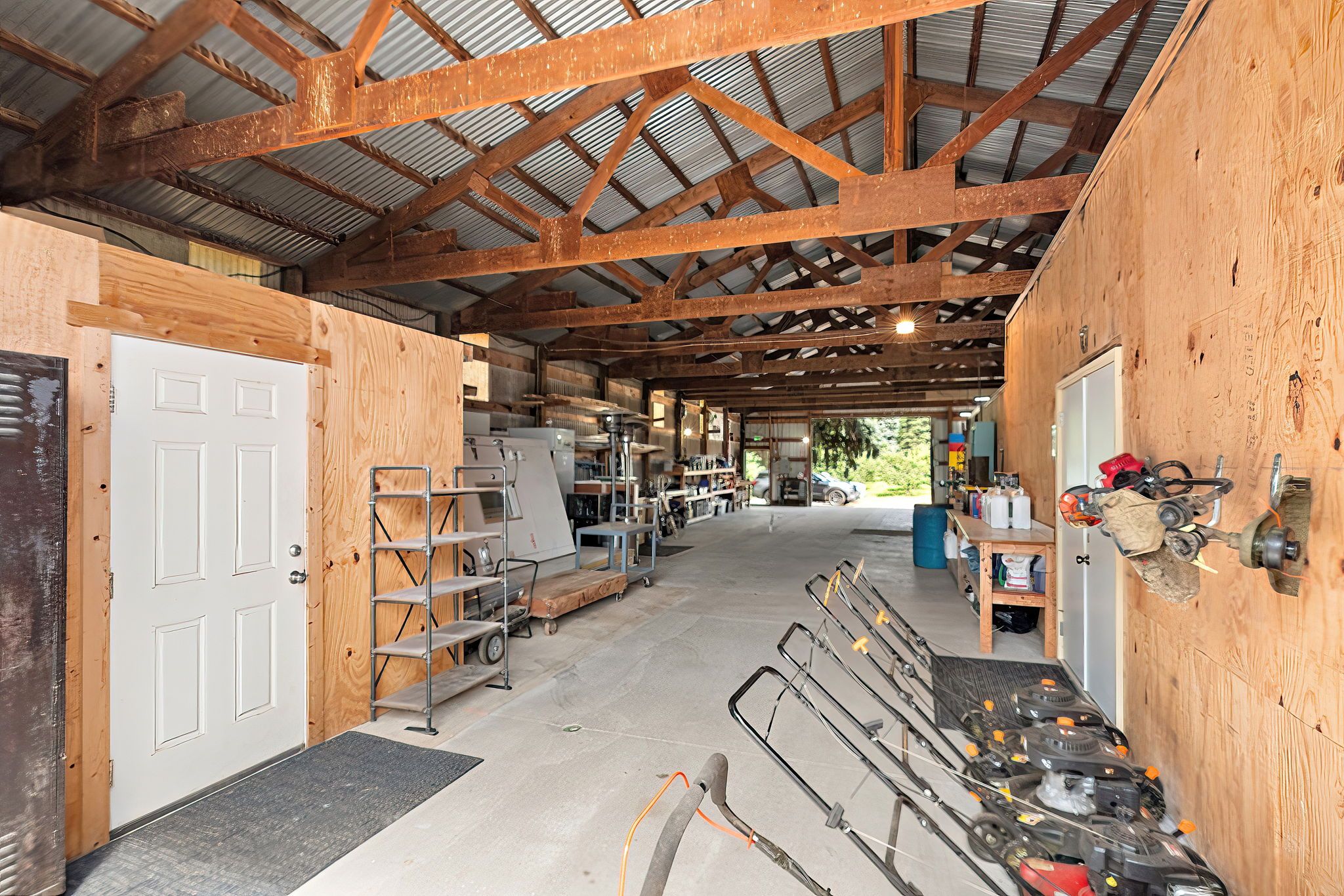 Interior of a spacious, rustic shed with a metal roof, wooden beams, and plywood walls, housing lawnmowers, tools, metal shelving, and various equipment.