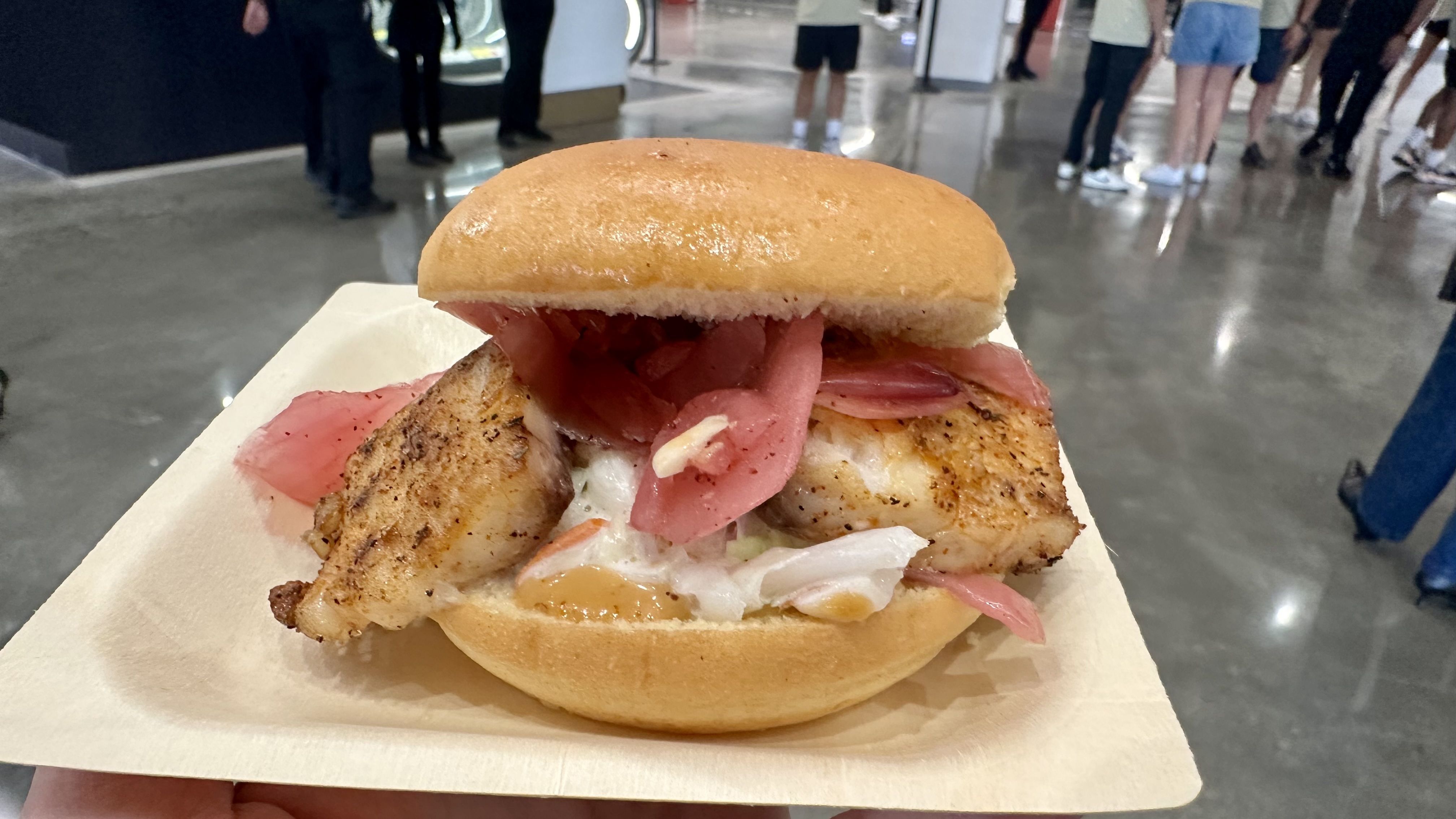 Grilled fish sandwich with pickled pink onions and creamy coleslaw on a soft bun, served on a paper tray in a busy indoor setting with people walking in the background.