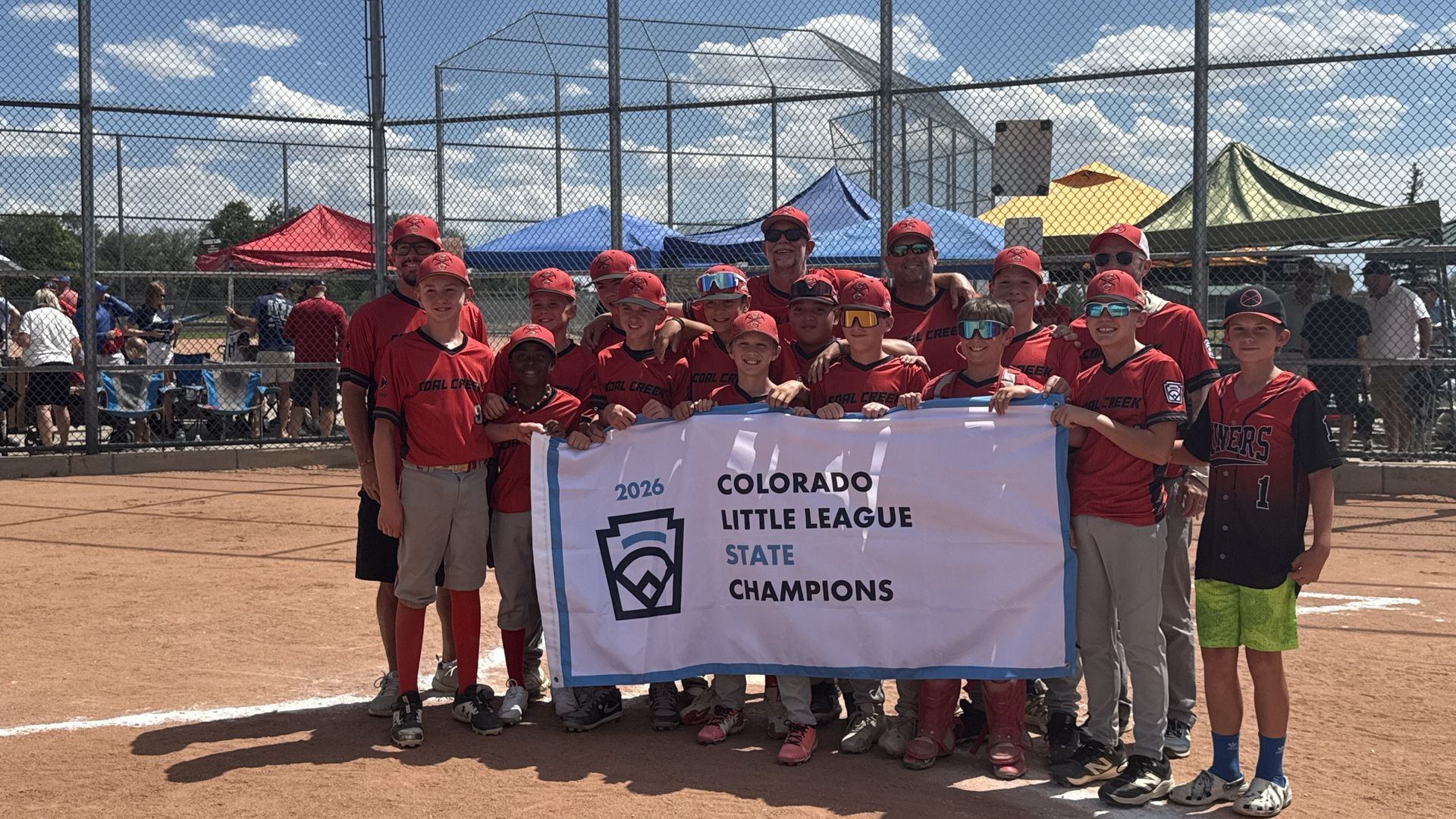Group of kids in red baseball uniforms posing with a banner reading "COLORADO LITTLE LEAGUE STATE CHAMPIONS" on a dirt field behind a chain-link fence, under a blue sky with clouds.