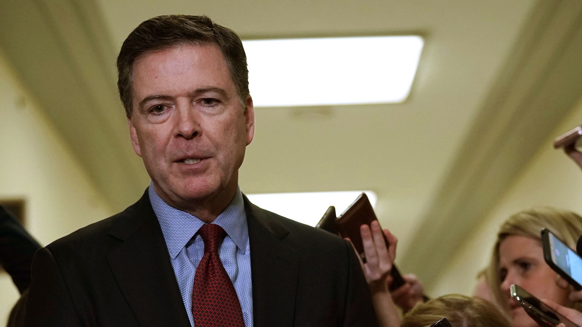 This is an image of James Comey looking down and speaking in a crowded hallway of reporters at Capitol Hill.