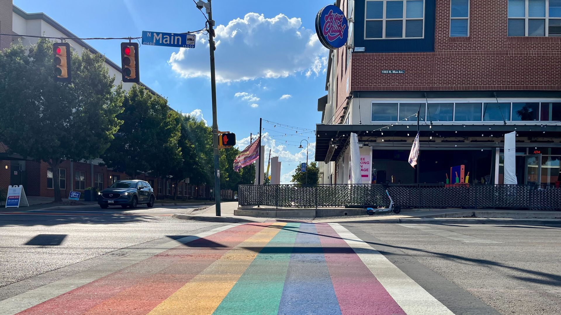 San Antonio Street intersection with rainbow-colored crosswalk under blue sky, traffic lights, and a sign for N Main. A building with a round "Jaycee's Ching" sign is on the corner.