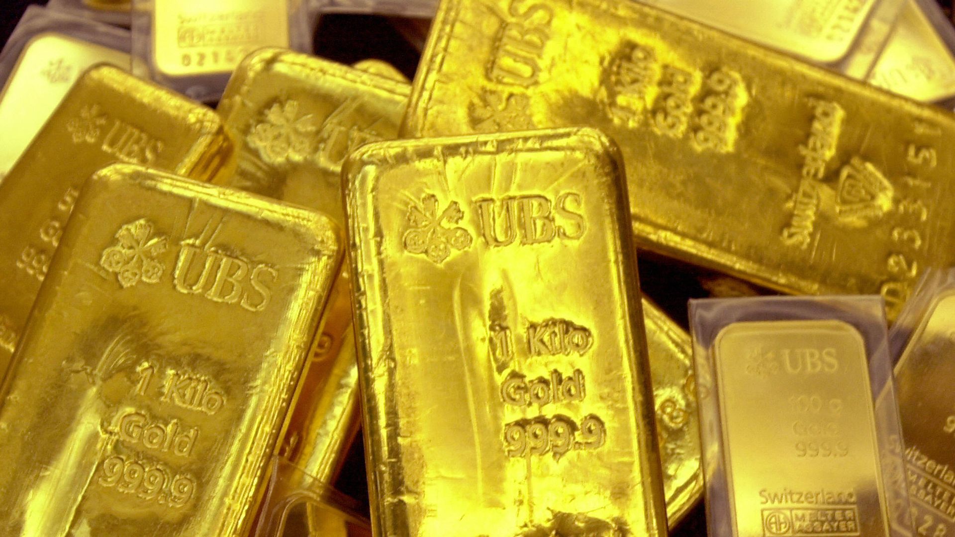 Pile of shiny gold bars marked with "UBS", weight, and purity details, some sealed in plastic, glowing yellow against a dark background.