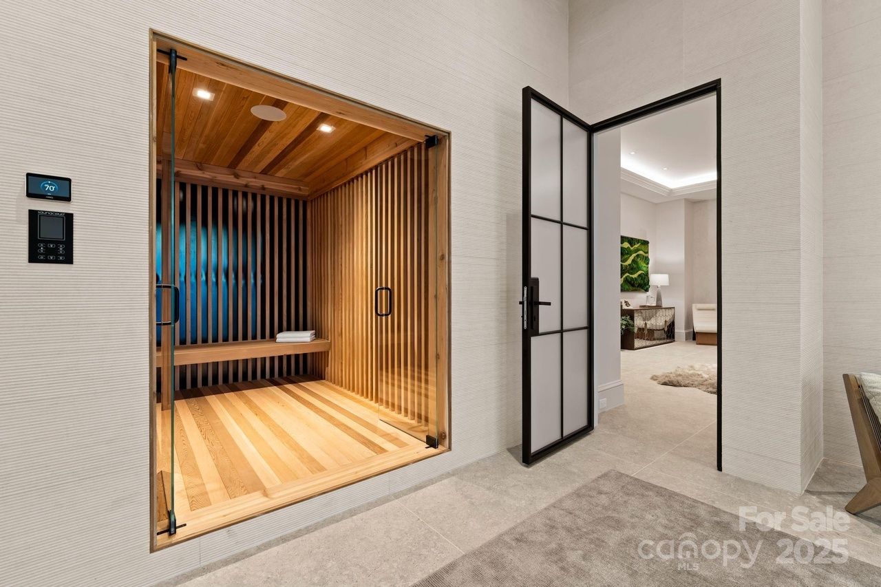 Modern room with light beige walls, a wooden sauna with glass doors and a wooden bench with towels inside, and a black-framed frosted glass door leading to a room with artwork and furniture.