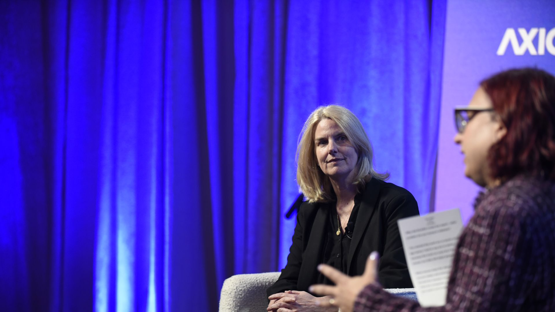 Panel discussion on a stage with blue curtains. A blonde woman in black sits center, facing forward; another person with glasses and reddish hair speaks on the right, with a paper visible.