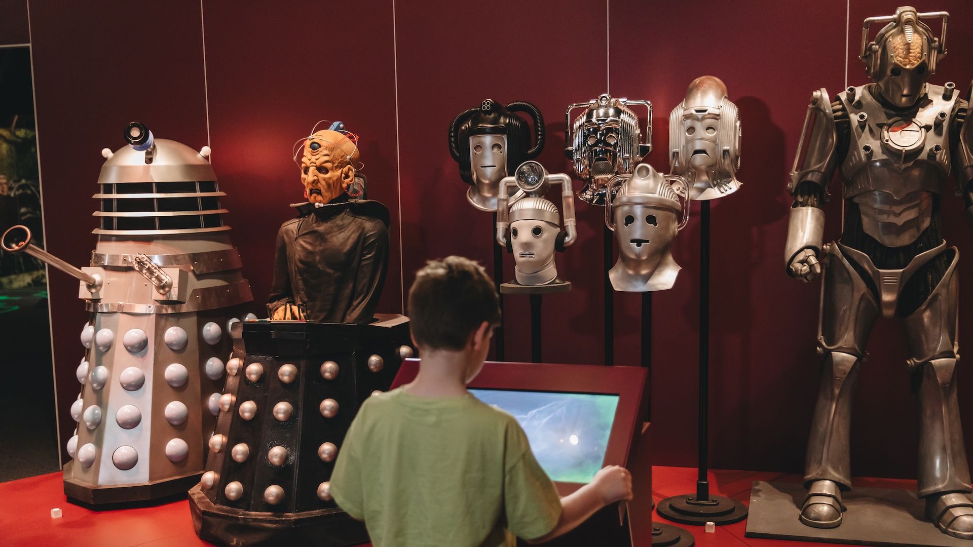 A boy stands at a kiosk in front of a museum exhibit of costumes from "Doctor Who" villains.