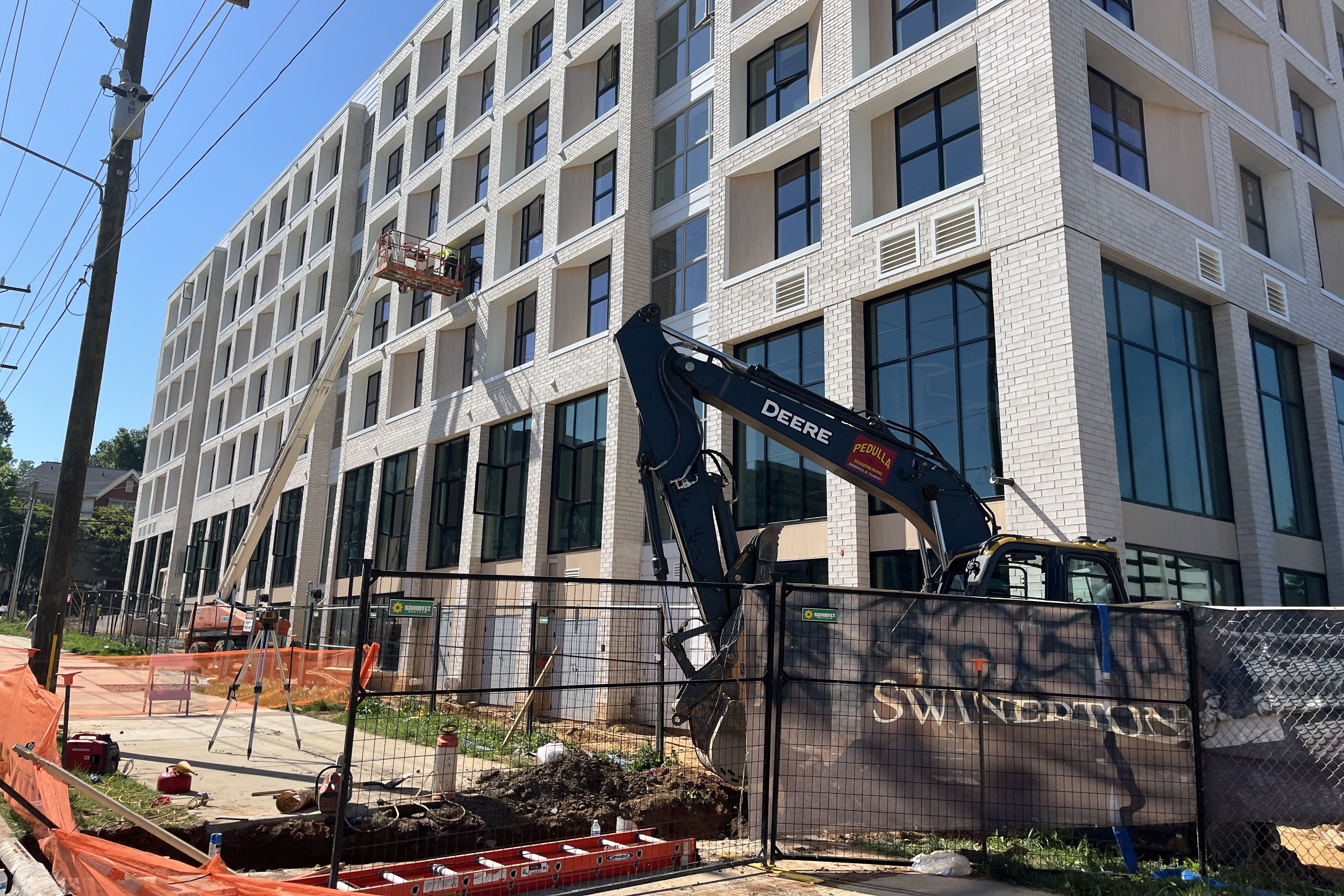 Construction site beside a white brick building with large glass windows. A worker on a raised platform inspects the facade while a Deere excavator sits nearby; orange safety fencing surrounds site.