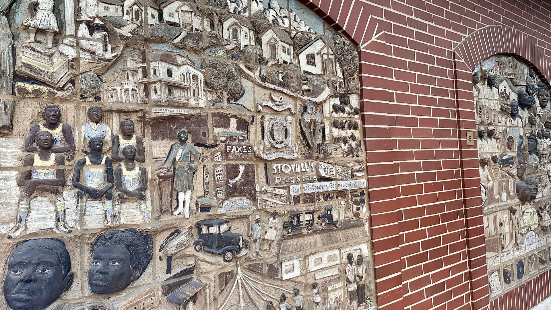 Relief mural on a brick wall featuring carved figures, historic buildings, vintage cars, and scenes including a group in sports uniforms and a drug store sign, with brown, beige, and gray tones.