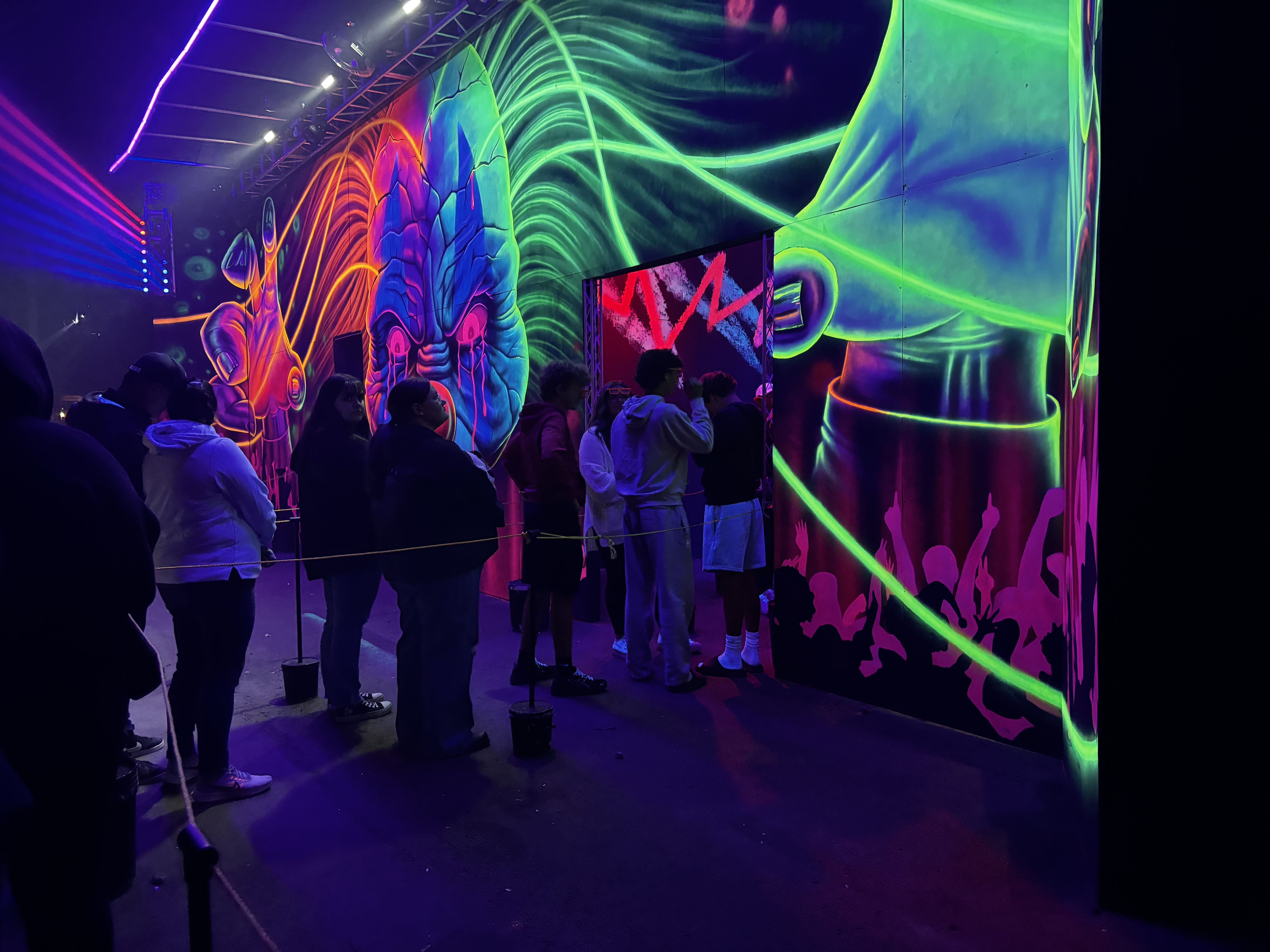 People lined up indoors by neon-lit walls with a large colorful mural of a blue face with red tears and green glowing hair, amid club-like purple and red lighting.