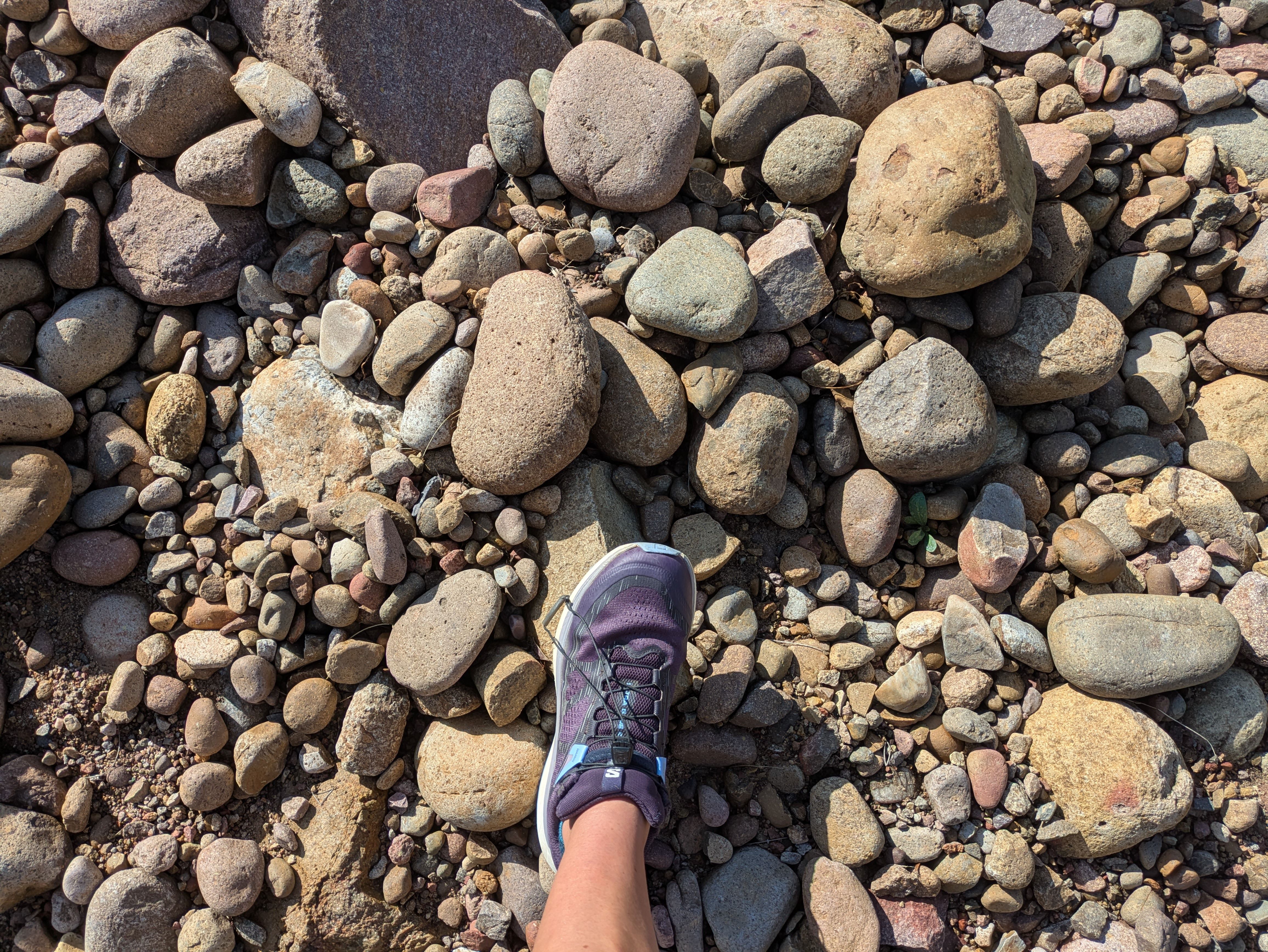 From a first-person view of a rocky ground covered with smooth, multicolored pebbles; a purple sneaker is visible at the bottom center.