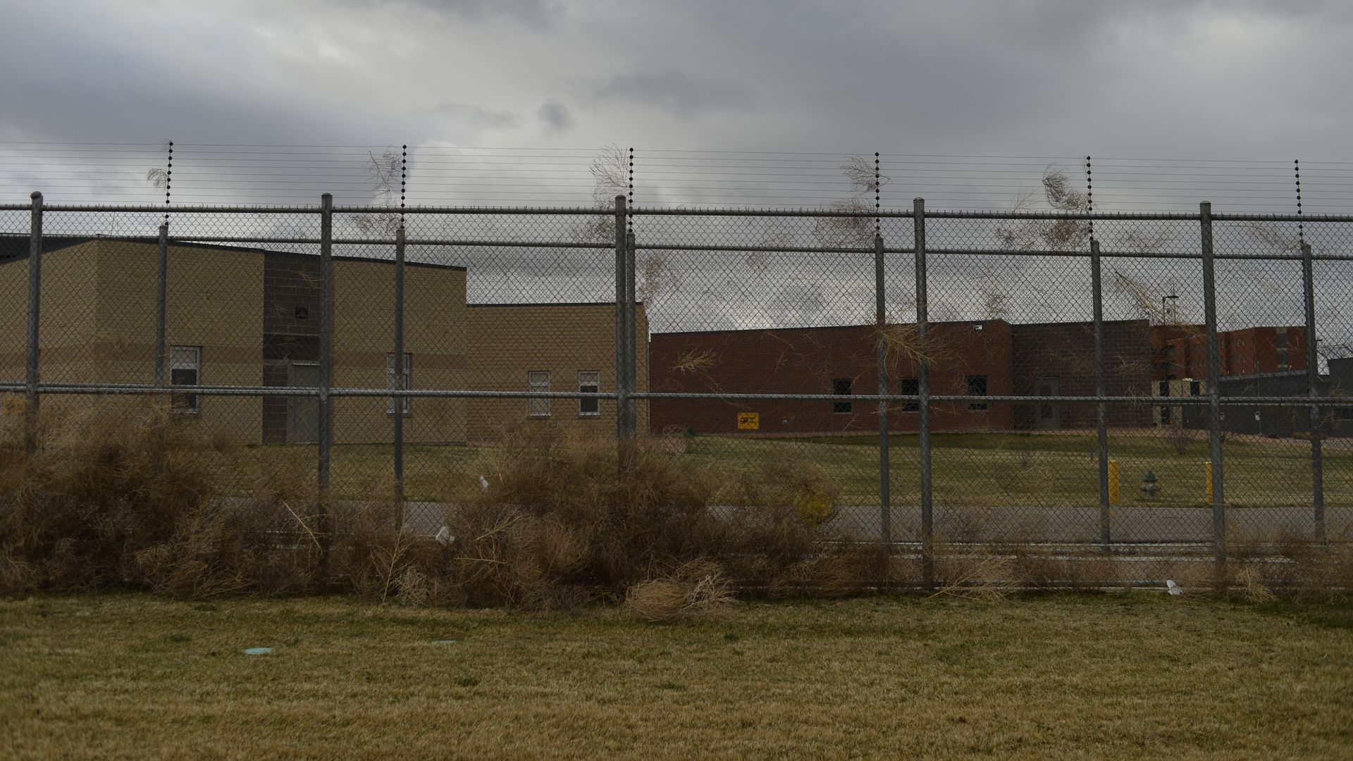 Cloudy sky over a tall barbed-wire fence and chain-link barrier, with tan and brown brick industrial buildings behind; dry shrubs in the foreground.