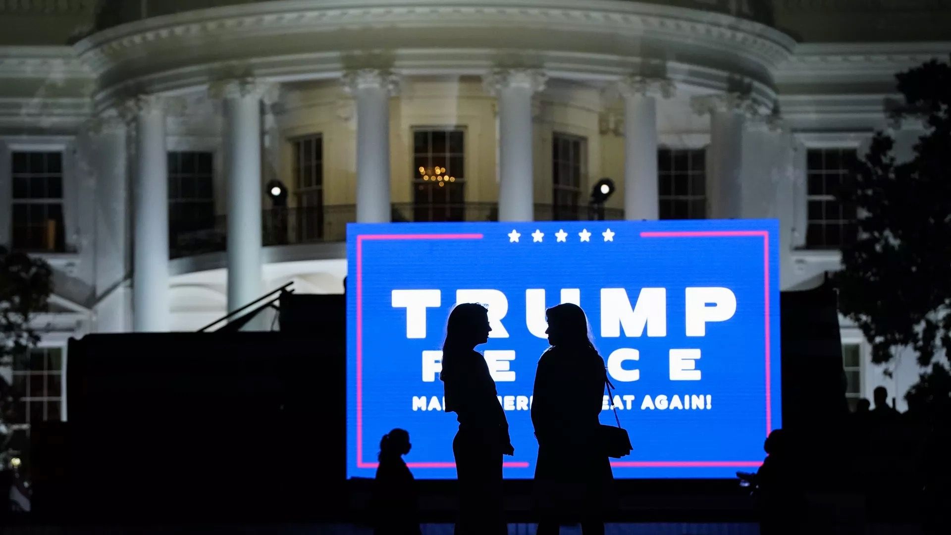 Two women are seen in silhouette standing in front of a Trump-Pence campign sign on the White House lawn.