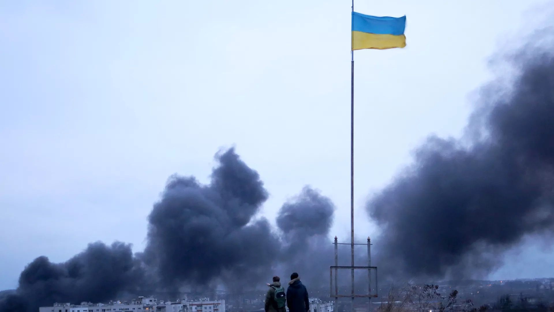 Onlookers view smoke from an airstrike as they stand before Ukraine's flag.
