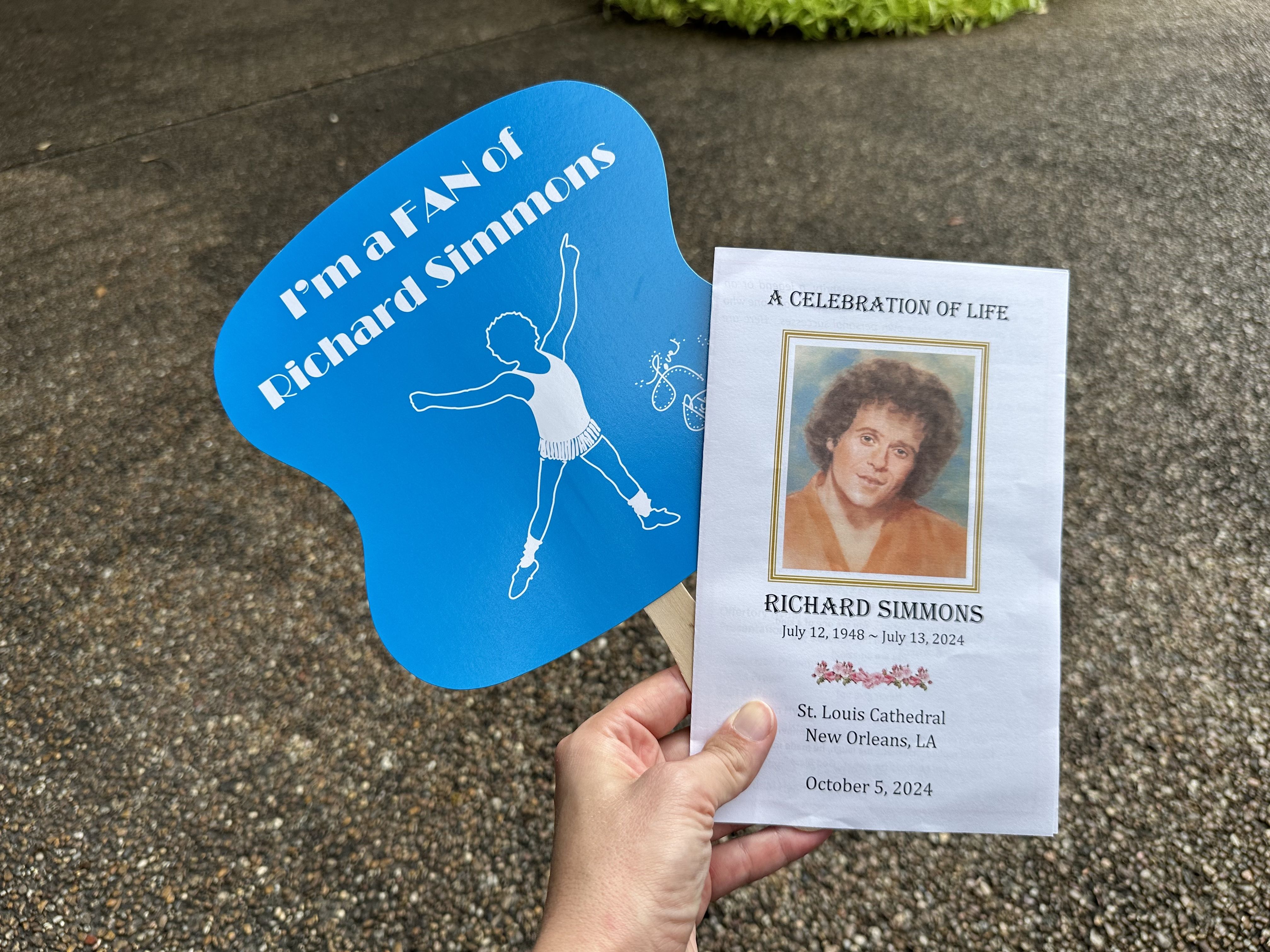 A photo of a paper fan that says "I'm a FAN of Richard Simmons" and a memorial service program being held up.