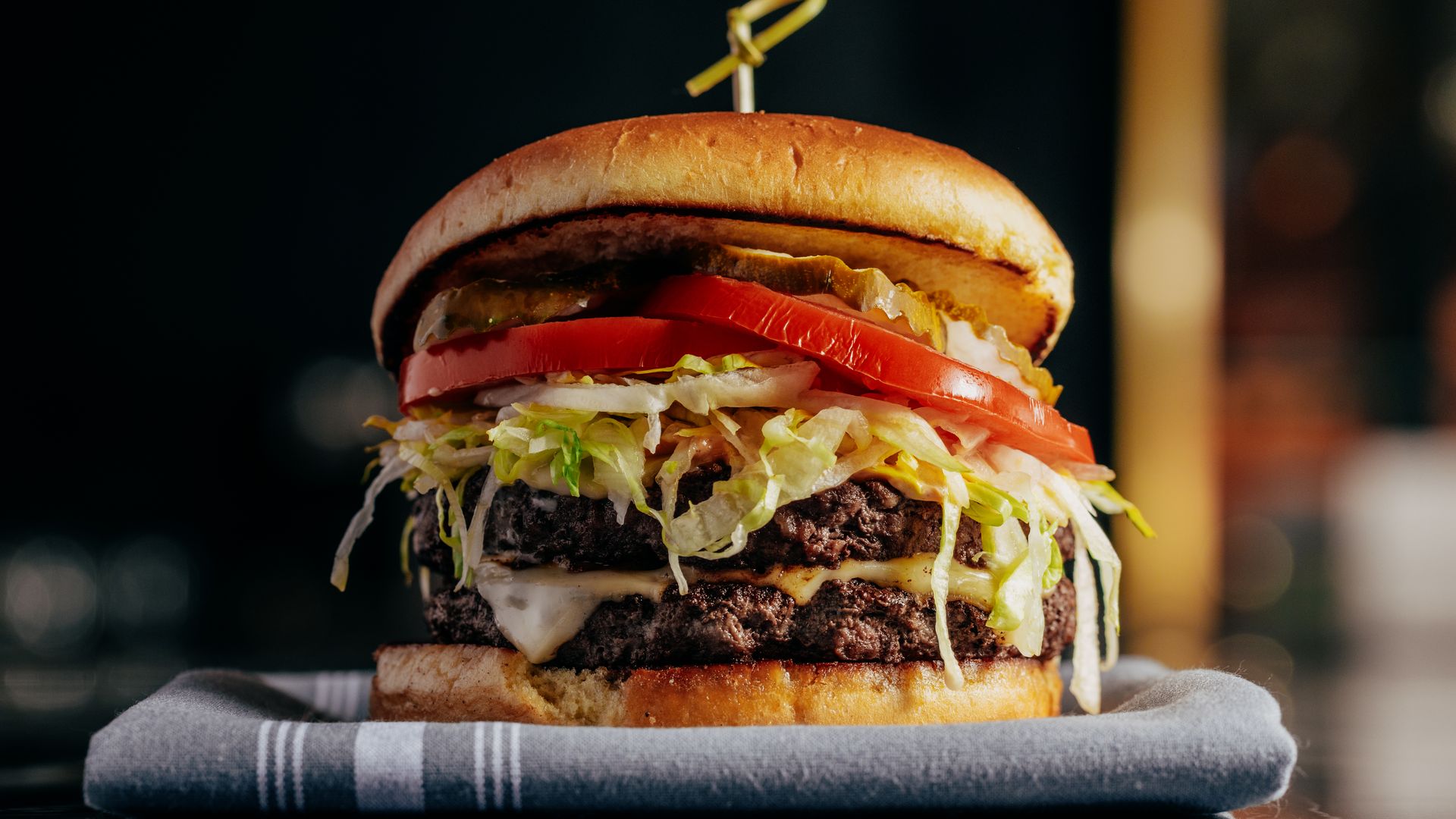 Close-up of a double cheeseburger with shredded lettuce, tomato slices, pickles, and melted cheese on a toasted bun, placed on a gray cloth against a dark blurred background.