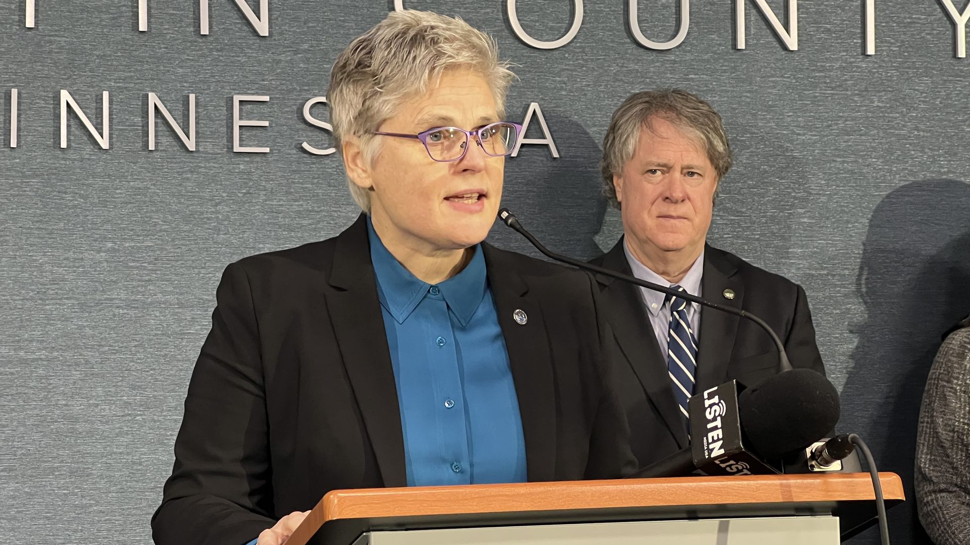 Person with short gray hair and purple glasses speaking at a podium with a microphone, wearing a black blazer and blue shirt, with a man in a suit and striped tie standing behind them.