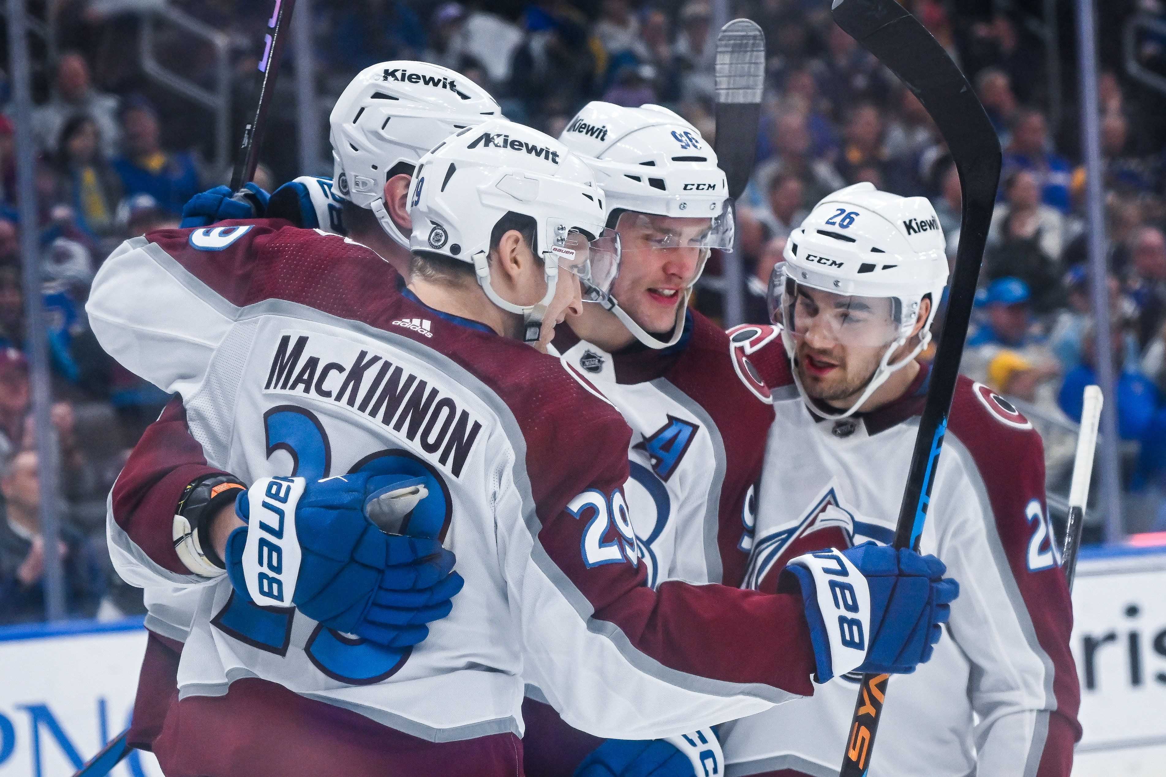 Four hockey players in white and burgundy jerseys, wearing white helmets, hug one another while celebrating a goal inside a packed arena.