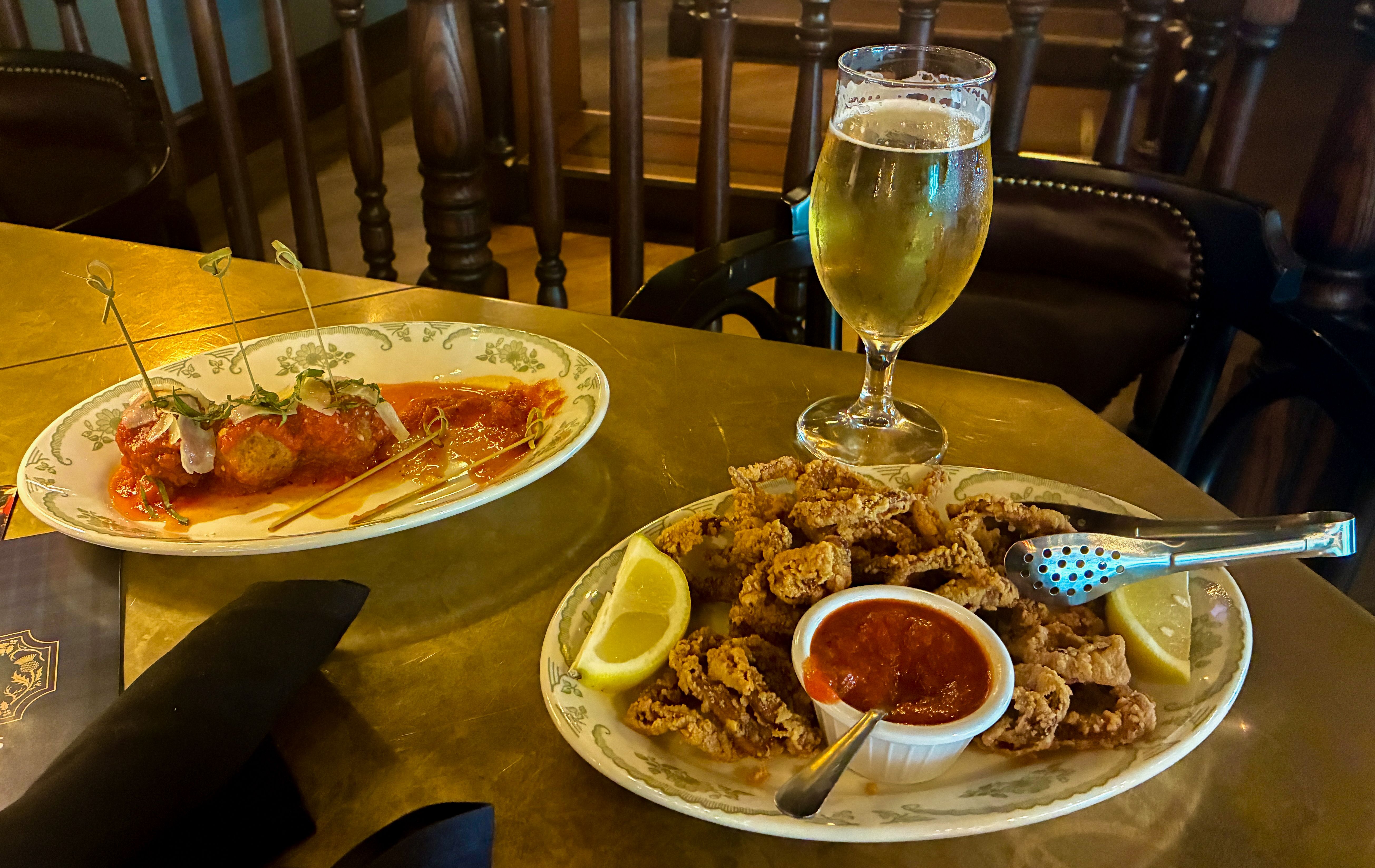 Plate of fried calamari bites with red sauce and lemon wedges on a patterned plate, a second plate of fried morsels with sauce, and a tall glass of pale beer; dark wooden chairs in the background.