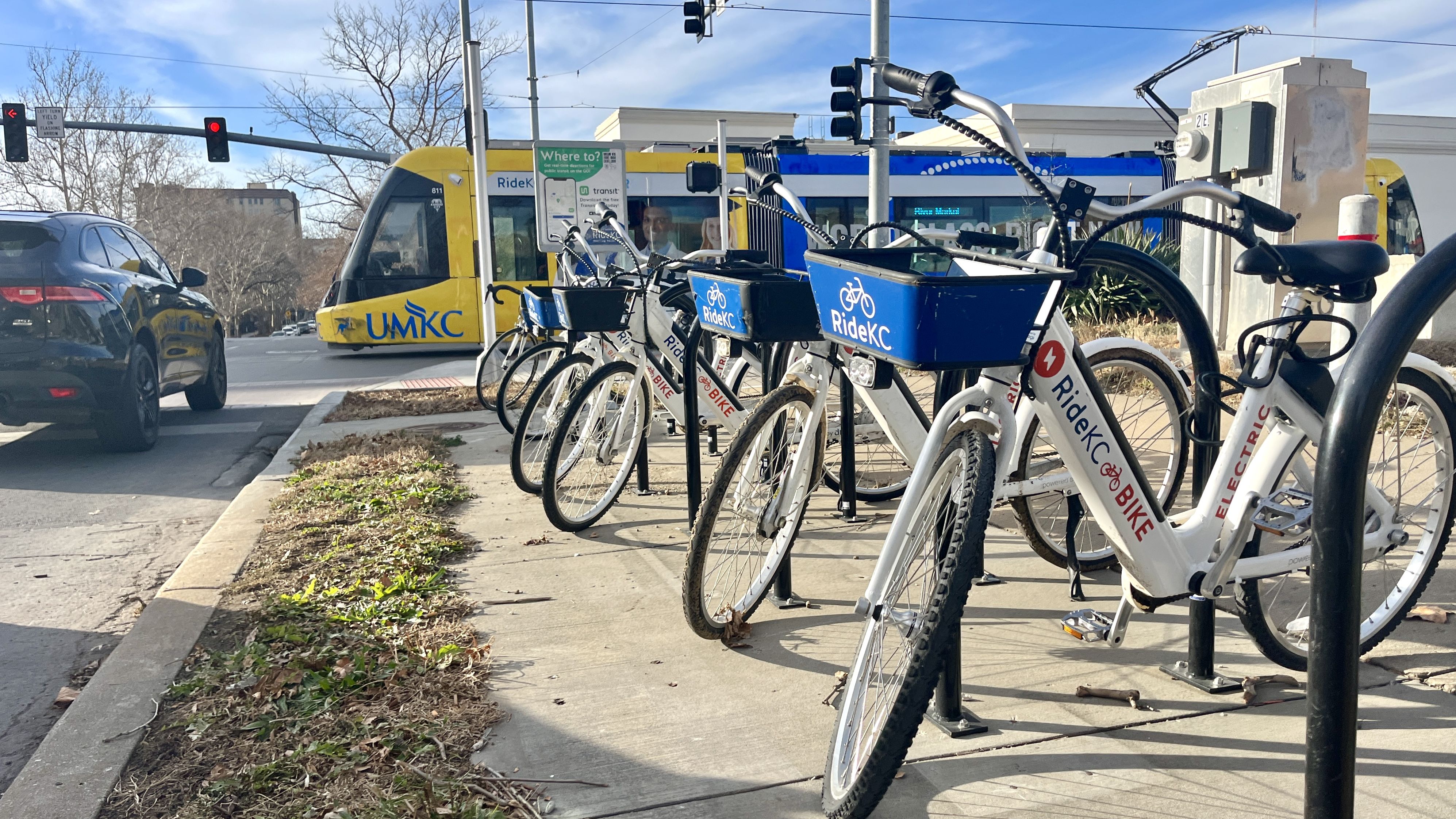 Row of white RideKC electric bike rentals with blue front baskets parked on a sidewalk near a street intersection, with a yellow and blue UMKC streetcar and a black car stopped at a red light.