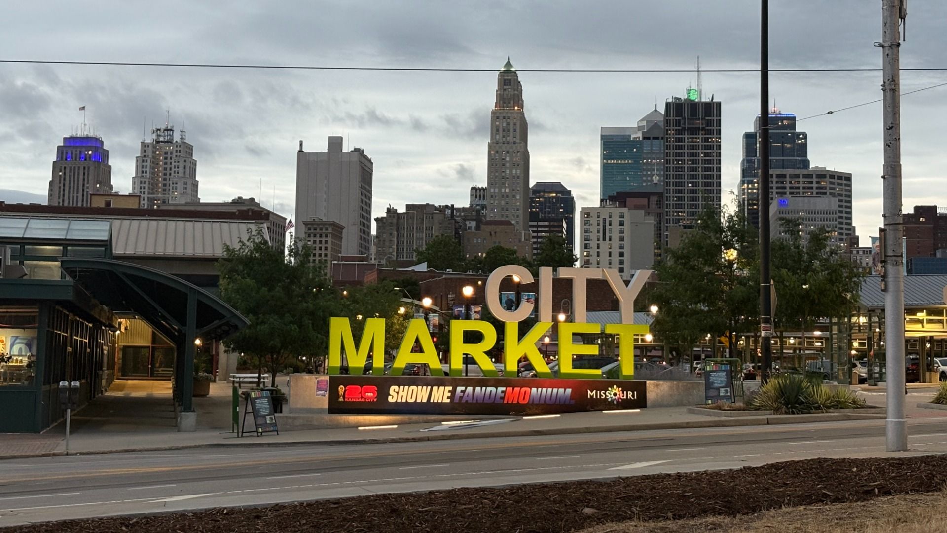 Wide view of a city market entrance with huge yellow letters spelling MARKET and gray CITY above, set against a downtown skyline of tall buildings on a cloudy day; a banner runs along the base.