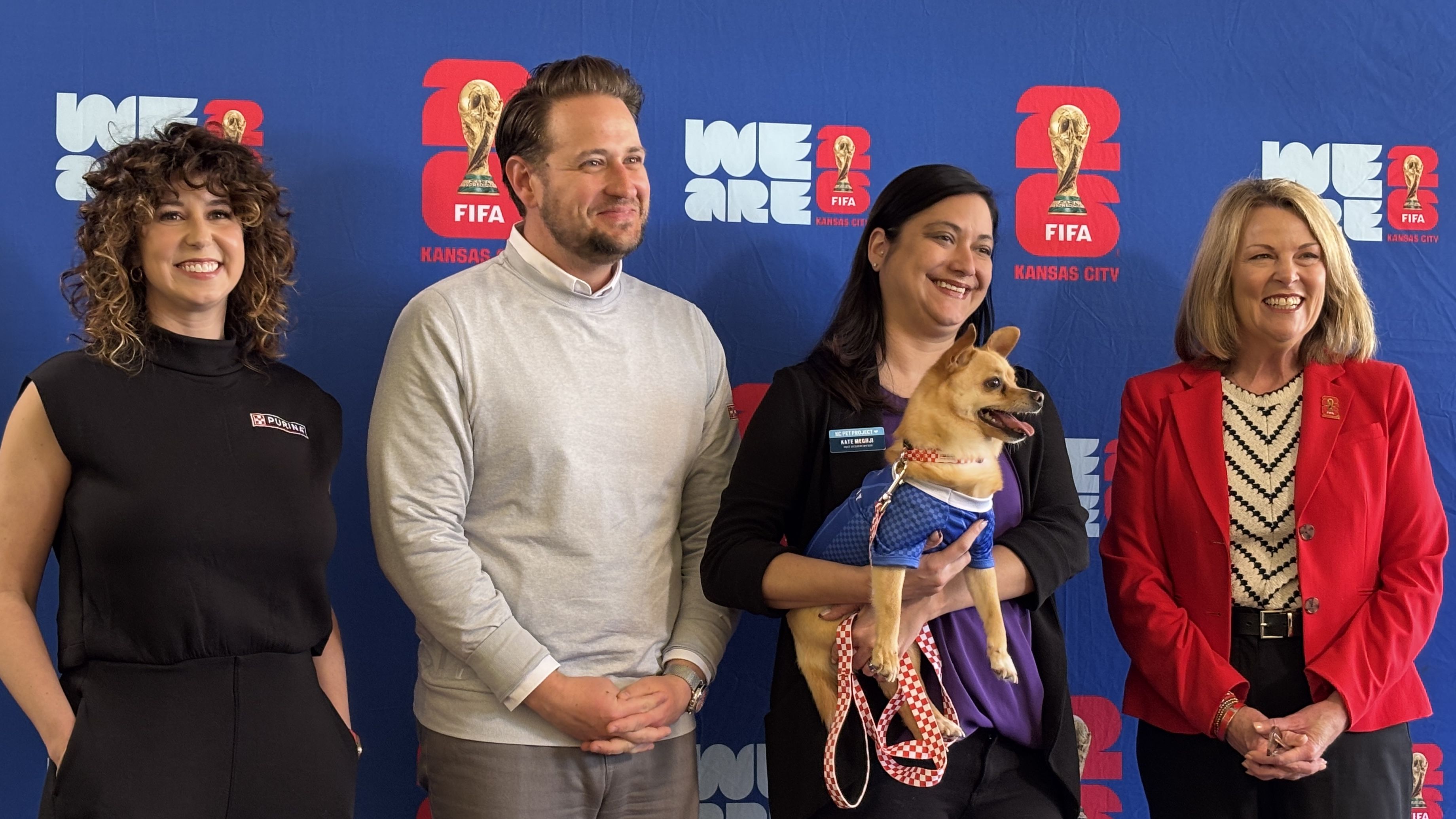 Four adults pose in front of a blue FIFA Kansas City backdrop; left to right: curly-haired woman in black, man in gray, woman with a small tan dog in a blue outfit, and a woman in a red blazer.