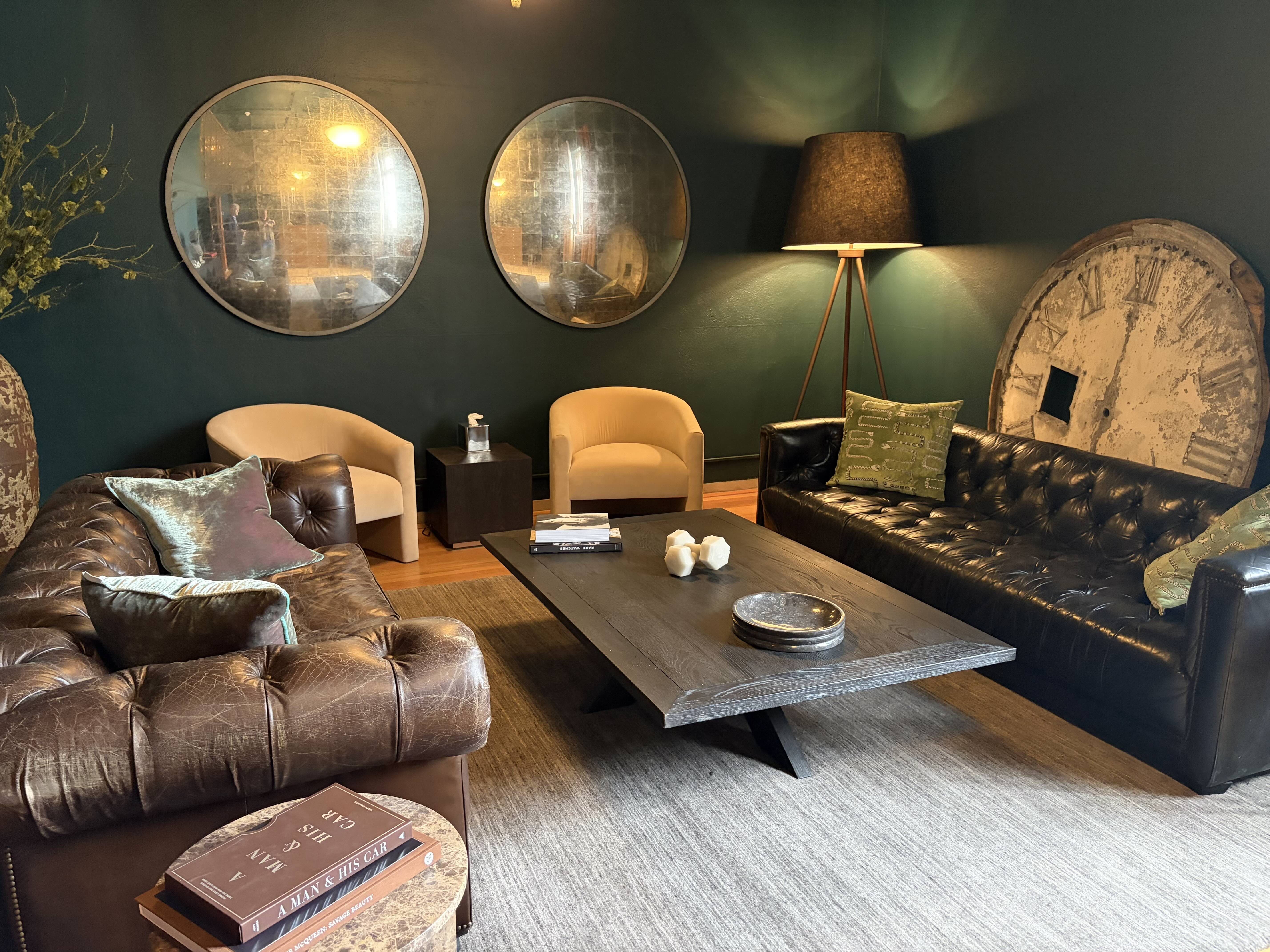 Cozy living room with dark teal walls, two large round mirrors, a tripod floor lamp, cream chairs, and a brown leather sofa. A rustic circular clock leans against the wall beside a coffee table.