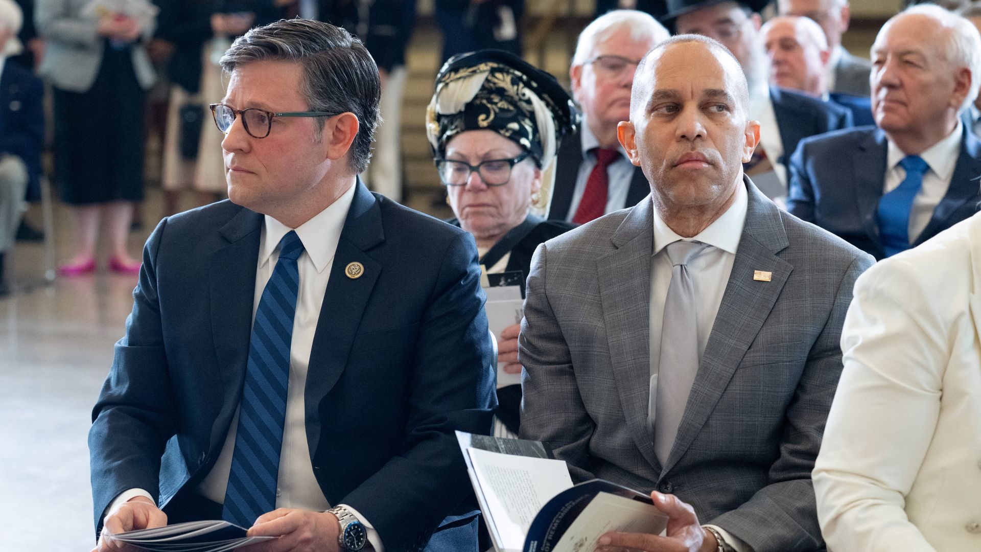 Two men in business suits sit at a formal event. Left man with glasses and a blue striped tie; right man in a gray suit holds a program. A woman wearing a patterned headscarf sits behind them.