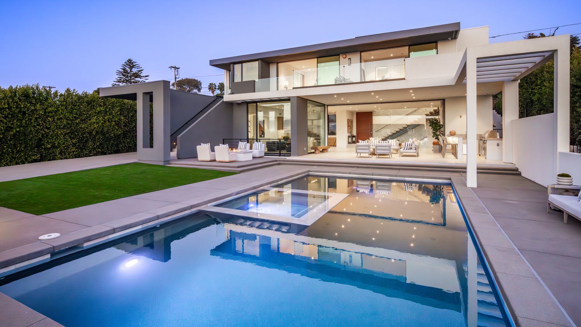 Modern two-story house with expansive glass walls, outdoor seating area, and a rectangular pool reflecting the lit interior. Lawn and tall hedges lining left; stairs and pergola on right.