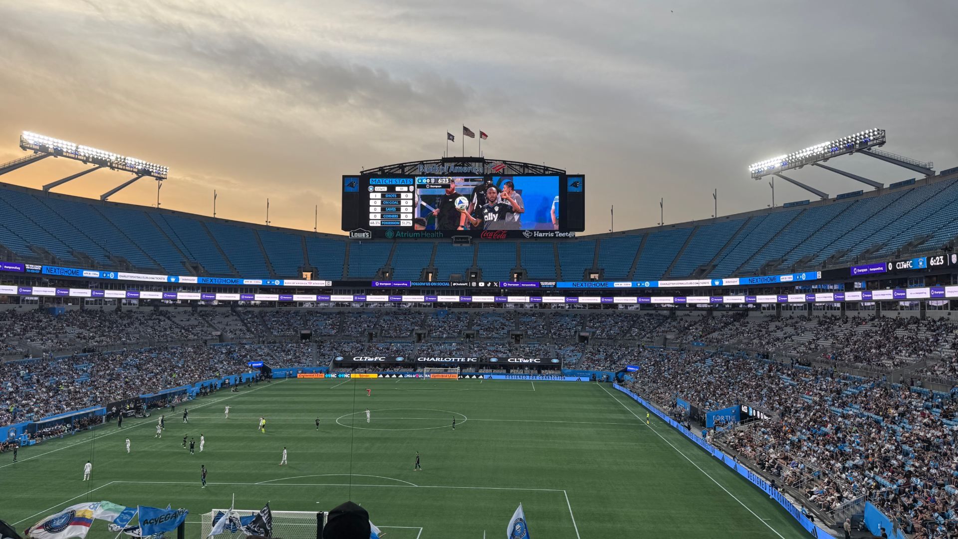 Wide view of a soccer stadium at dusk, with a big central video scoreboard, bright floodlights, and blue seating. Players on a green pitch while crowds fill the stands.