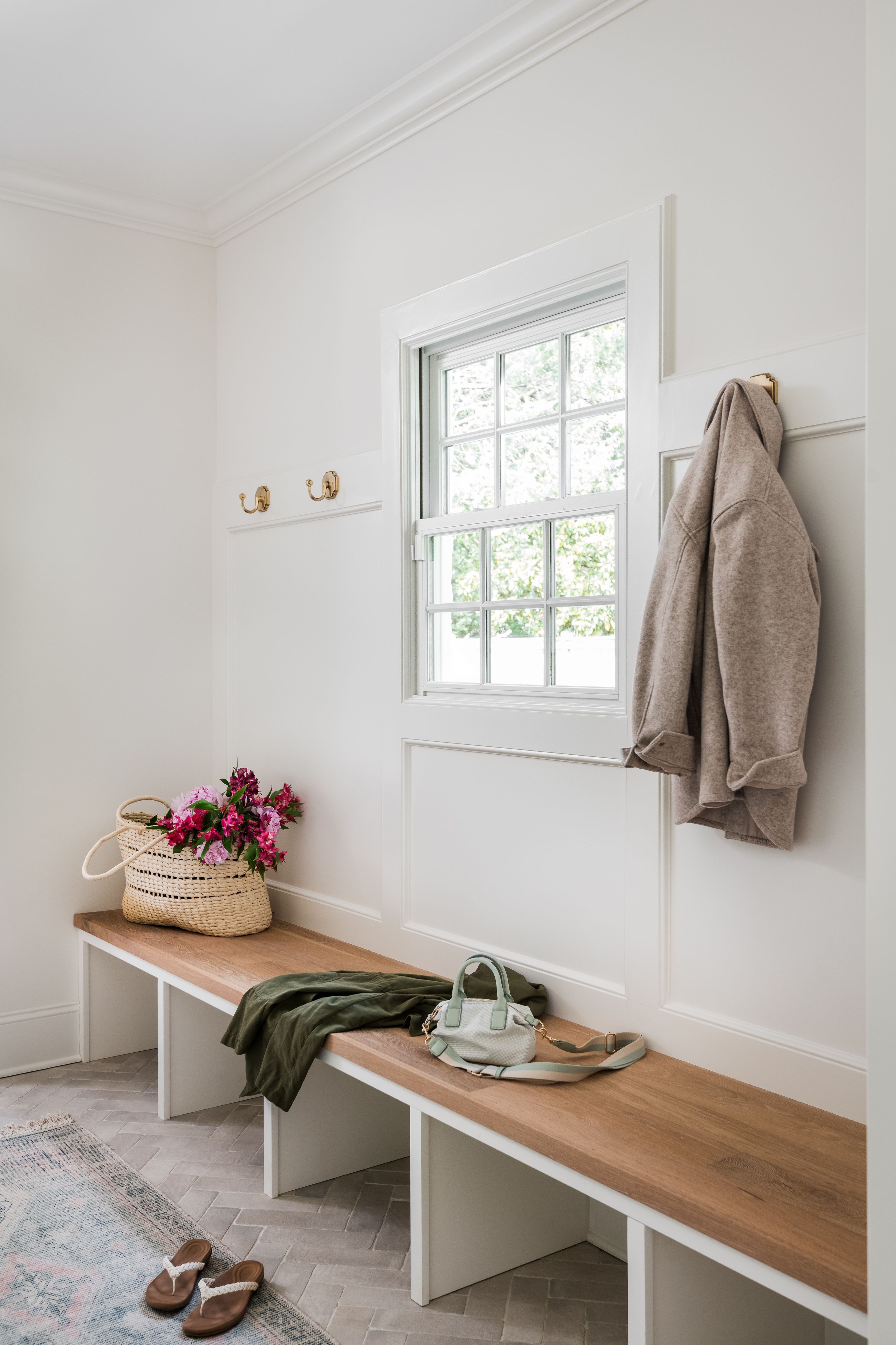 Minimalist entryway with white walls, wooden bench holding a wicker basket with pink flowers, green jacket, and small mint handbag; beige jacket hangs on gold hooks by window; sandals on floor.