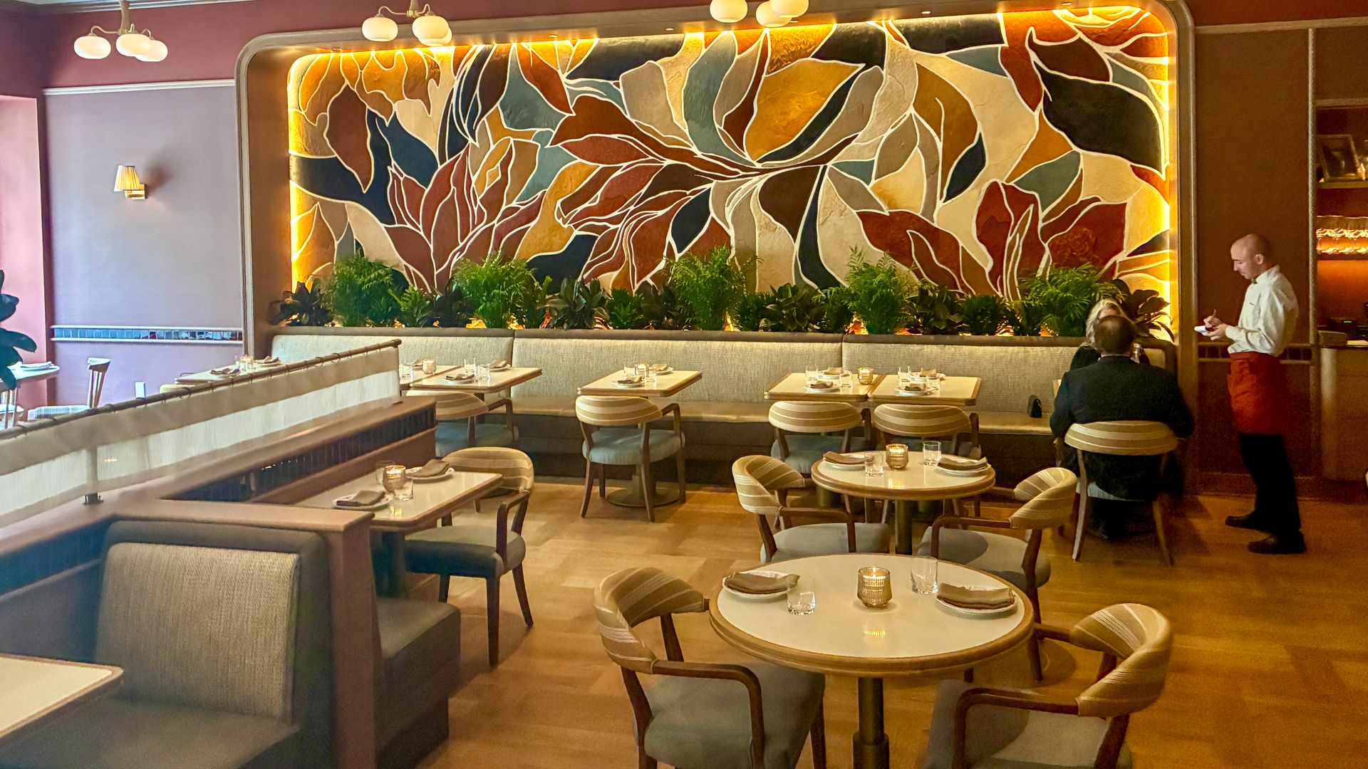 Upscale restaurant dining room with a large colorful leaf mosaic wall, warm backlit lighting, row of plants along a divider, and round tables set for guests; a server stands on the right.