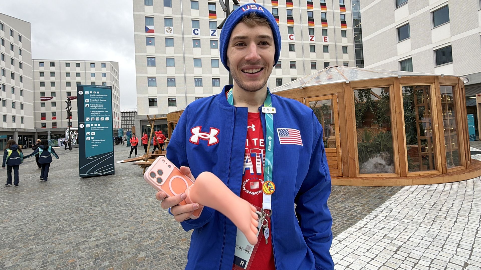 Smiling man in blue USA jacket and beanie holds a smartphone with a peach-colored case shaped like a foot, standing in a plaza with modern white buildings and a wooden structure behind him.