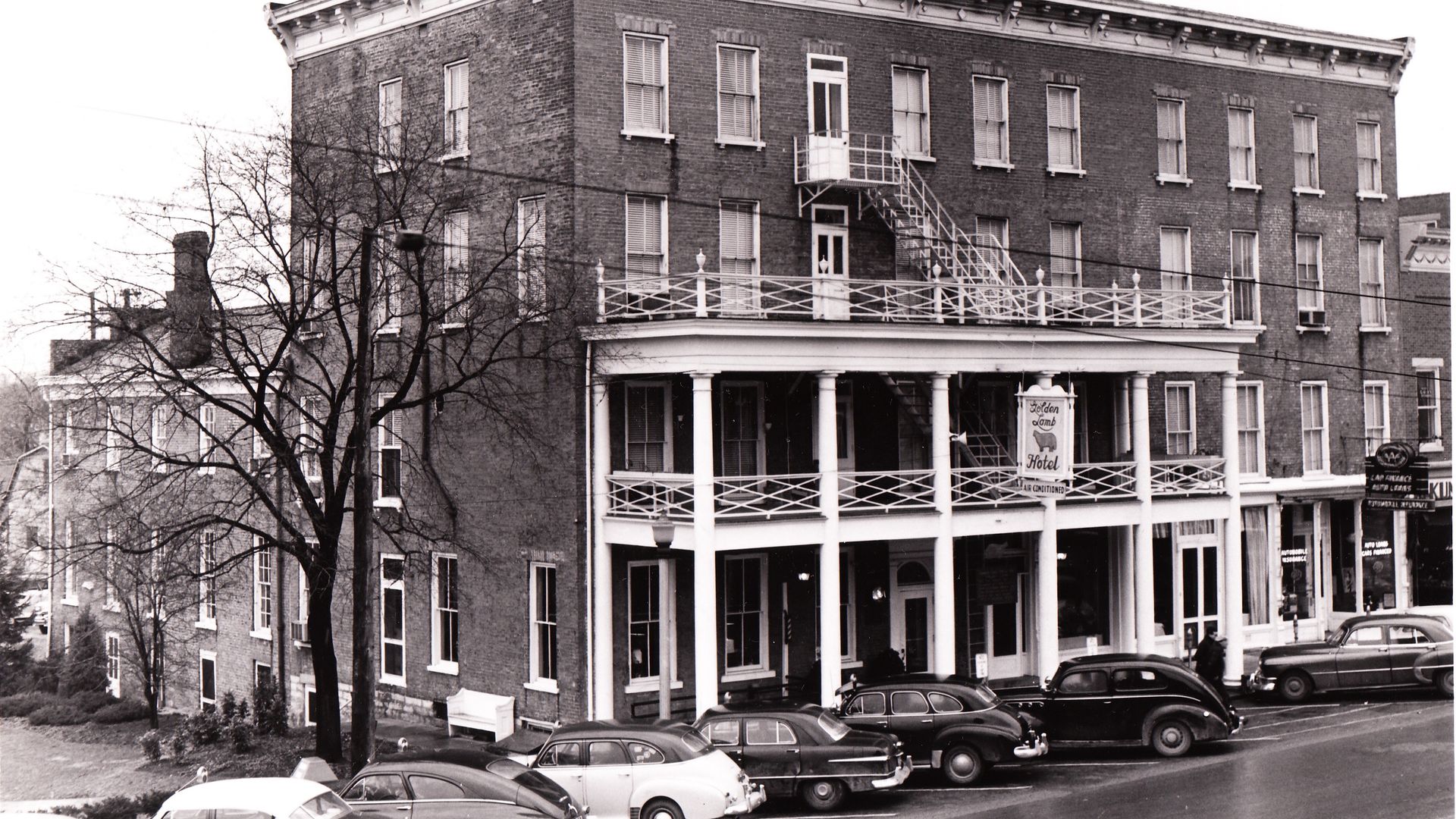 Black-and-white street scene of a brick hotel with ornate cornice, fire escape, and a row of vintage cars parked along the curb; a sign reads "Golden Lamb Hotel".
