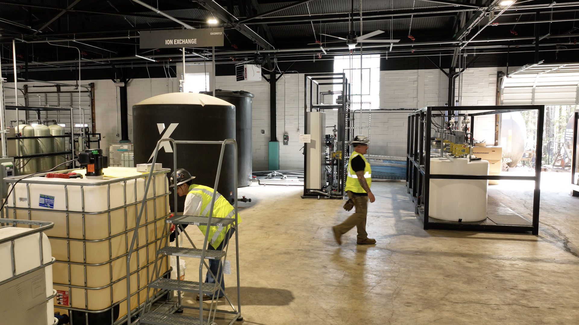 Industrial lab with a large black cylindrical tank bearing a white logo; overhead sign reads "ION EXCHANGE." Two workers in high-visibility vests and hard hats walk among pipes and tanks.