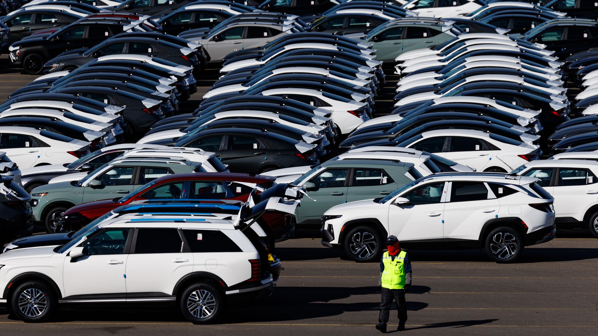 Rows of cars lined up at a Korean port, awaiting shipment. One worker, dressed in a yellow vest, stands in the foreground.