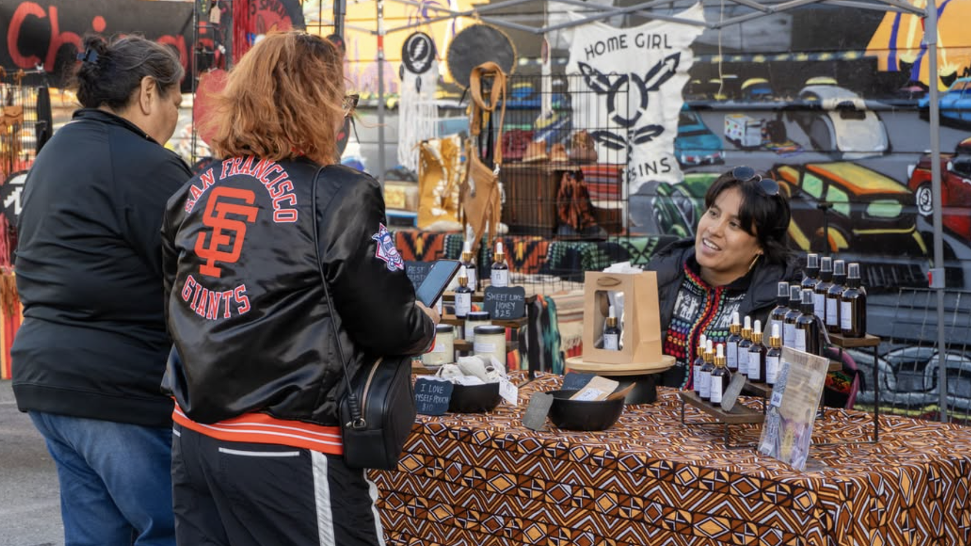 Two women at an outdoor market stall: one wearing a San Francisco Giants jacket chats with a vendor beside a table of bottles on a geometric cloth, with a colorful mural in the background.