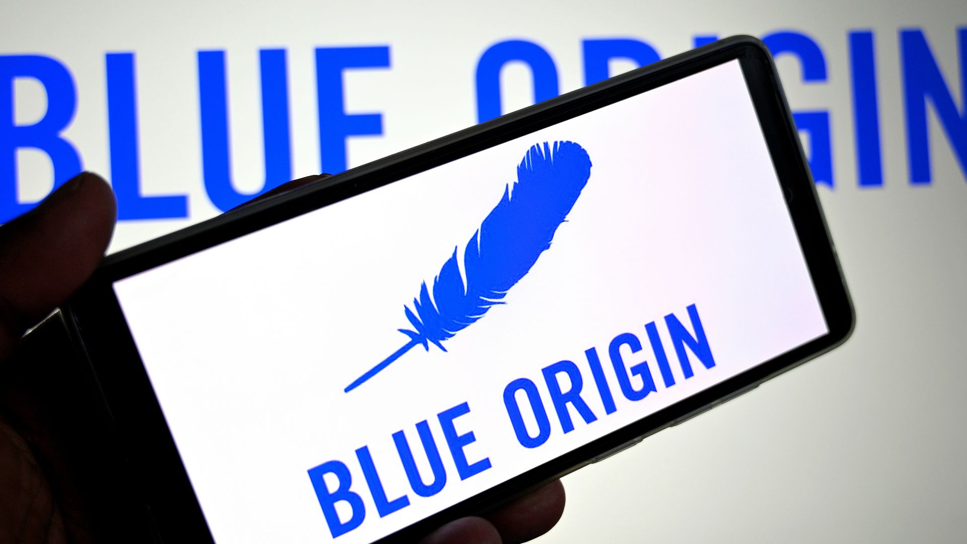 A smartphone in the foreground shows the Blue Origin logo, with Blue Origin written on the wall behind the phone as well.