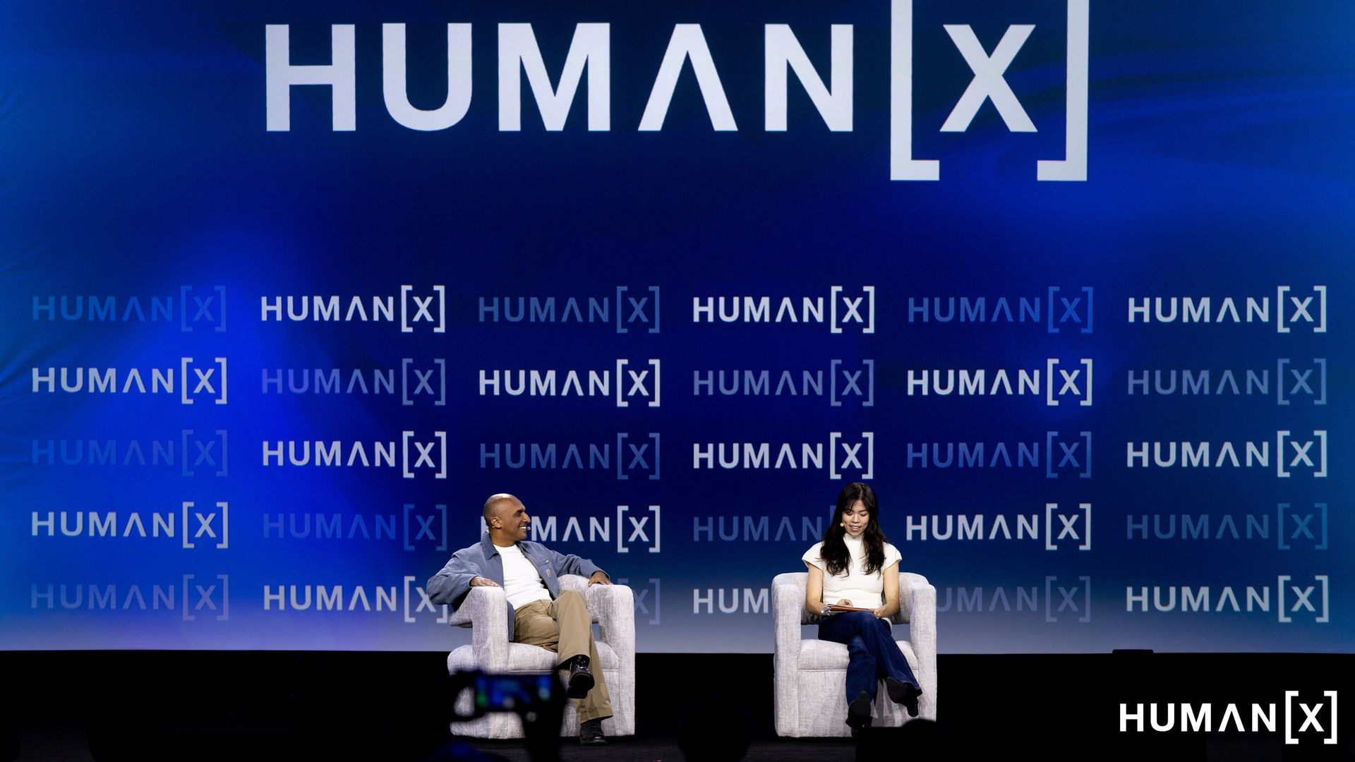 Blue gradient stage with a large 'HUMAN[x]' logo. Two people sit in white chairs; a man in a light jacket and beige pants chats with a woman in a white top and dark jeans, while repeated branding appears on the backdrop.