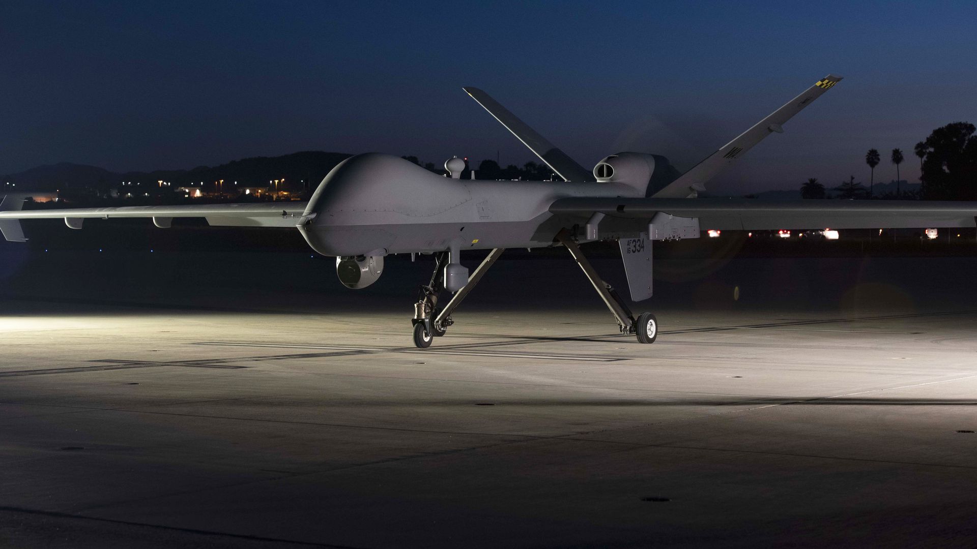 A large drone sits on a runway during dusk. Town lights can be seen in the background.