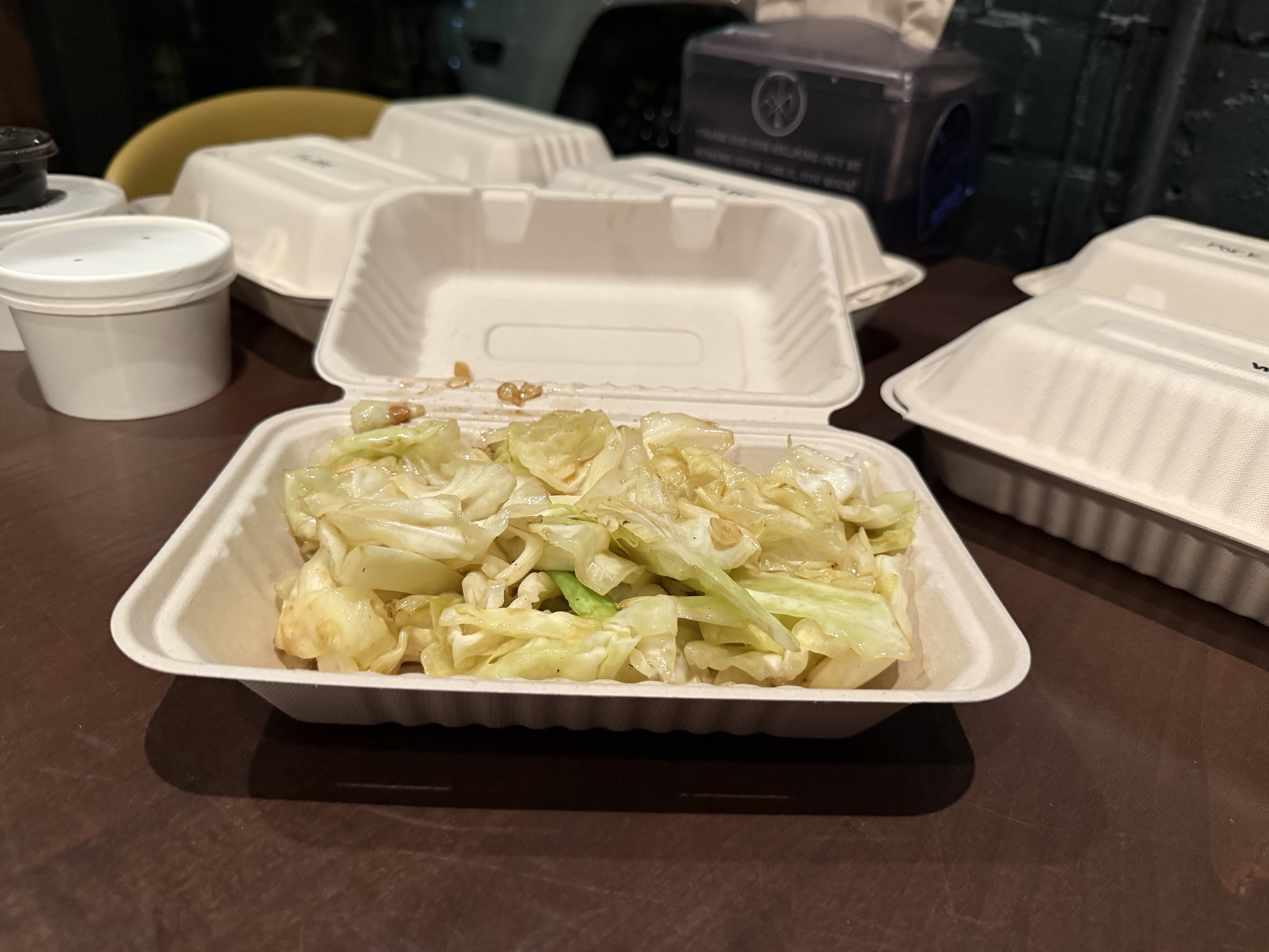 Takeout container on a table filled with cooked cabbage seasoned with garlic and sauce, surrounded by other closed white takeout containers and a white cup.