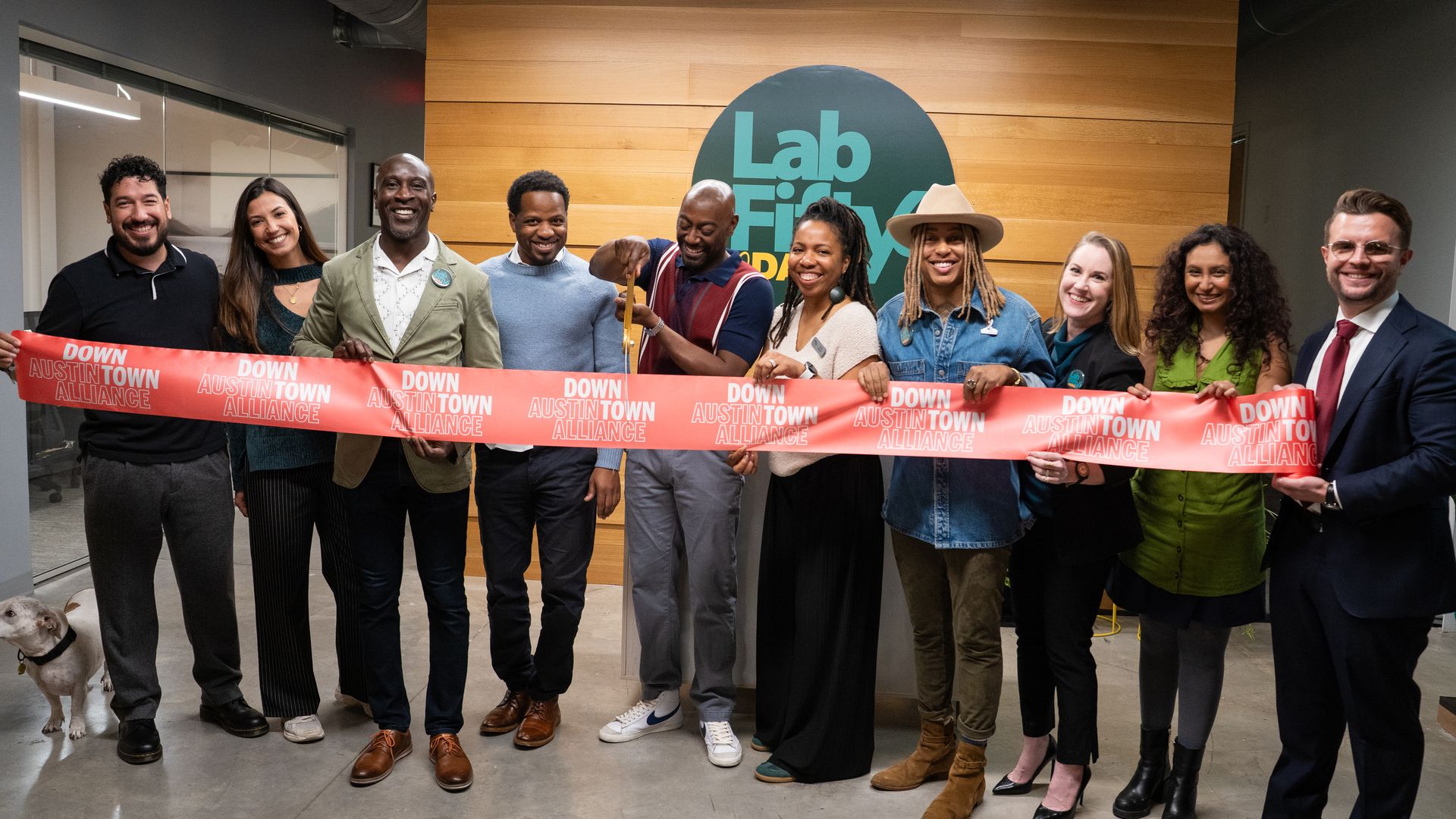 Group of nine diverse people smiling and holding a red ribbon that reads "Down AustinTown Alliance" in an office with wood wall and "Lab Finly" logo as one person cuts the ribbon.