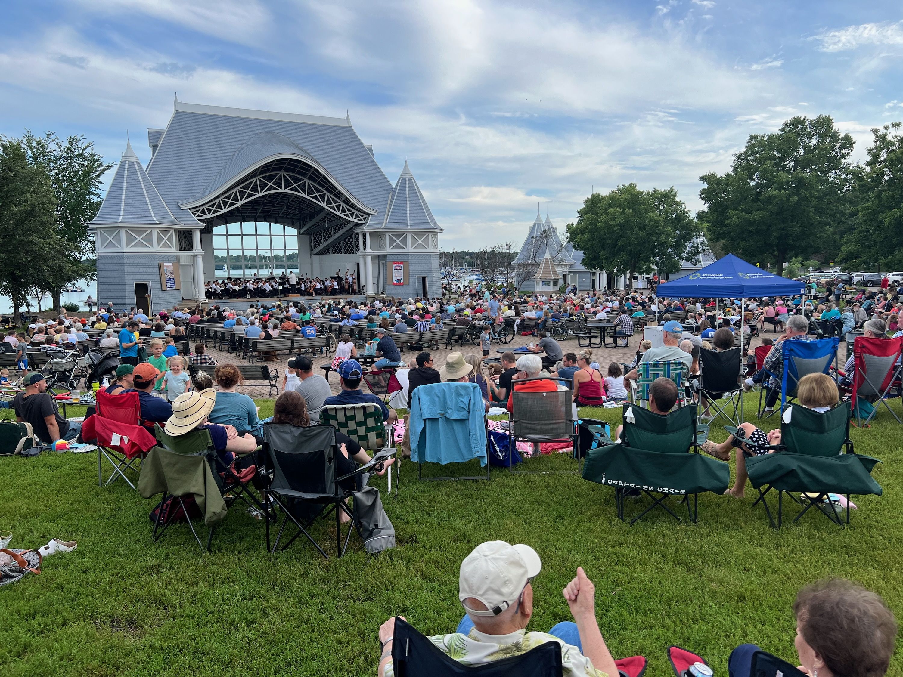 Outdoor concert at a lakeside park. A grand gray pavilion stage hosts an orchestra while crowds sit on benches and lawn chairs; blue canopies, trees, and a bright sky surround the scene.