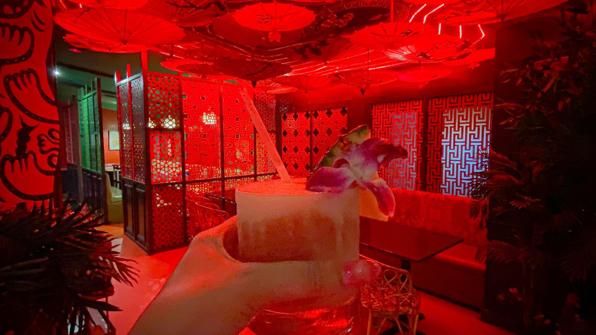 A light-colored cocktail is held in front of an empty room illuminated with red neon lights and decorated with hanging paper umbrellas.