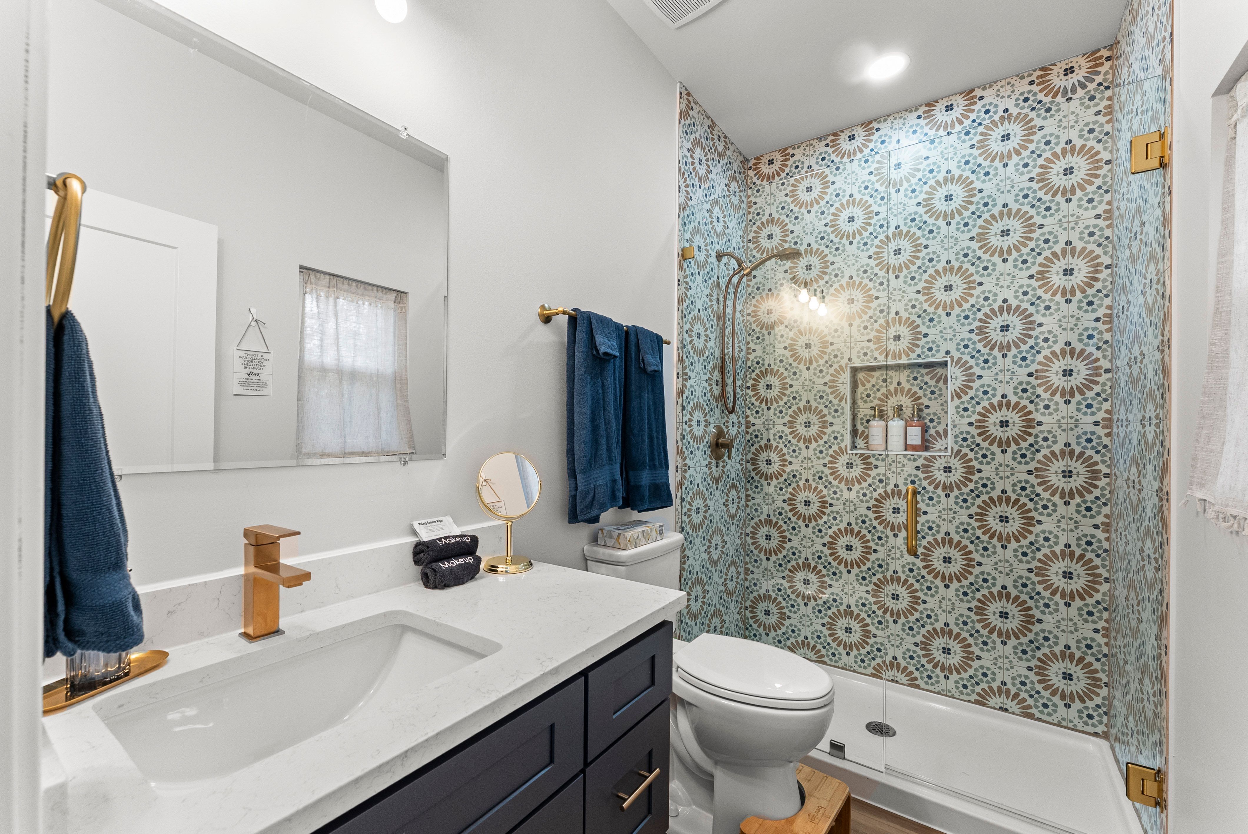 Bathroom with a white countertop and navy blue cabinets, gold faucet and fixtures, blue towels, and a shower with patterned teal, brown, and white tiles and gold hardware.